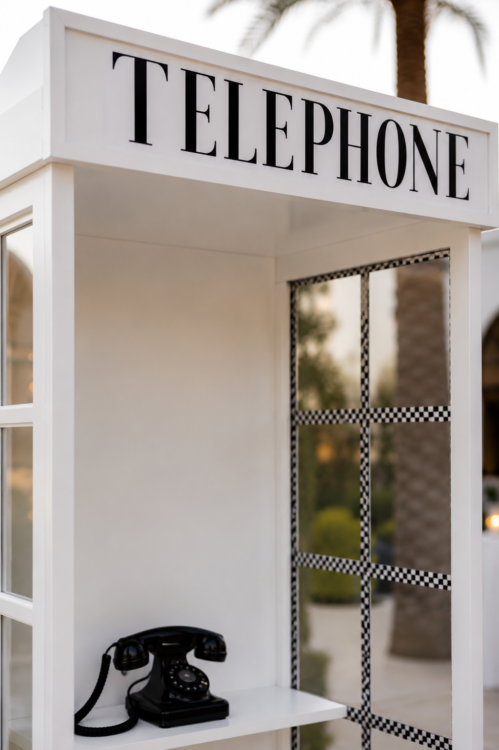 A vintage black rotary telephone inside a white telephone booth with a large sign reading 'TELEPHONE' on top, set outdoors with palm trees in the background.