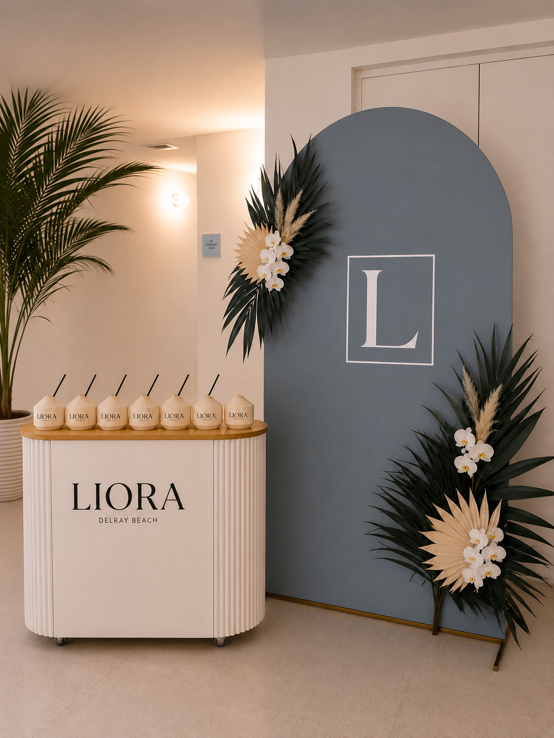 Decorative setup at Liora Delray Beach with a blue backdrop, large white 'L' logo, white flowers, palm fronds, and a white counter with 'LIORA DELRAY BEACH' and multiple coconut-shaped containers.