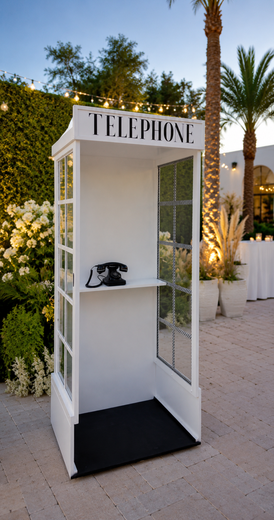 An outdoor white telephone booth with a vintage black rotary phone inside, decorated with a large 'TELEPHONE' sign on top, set in a garden area at dusk.