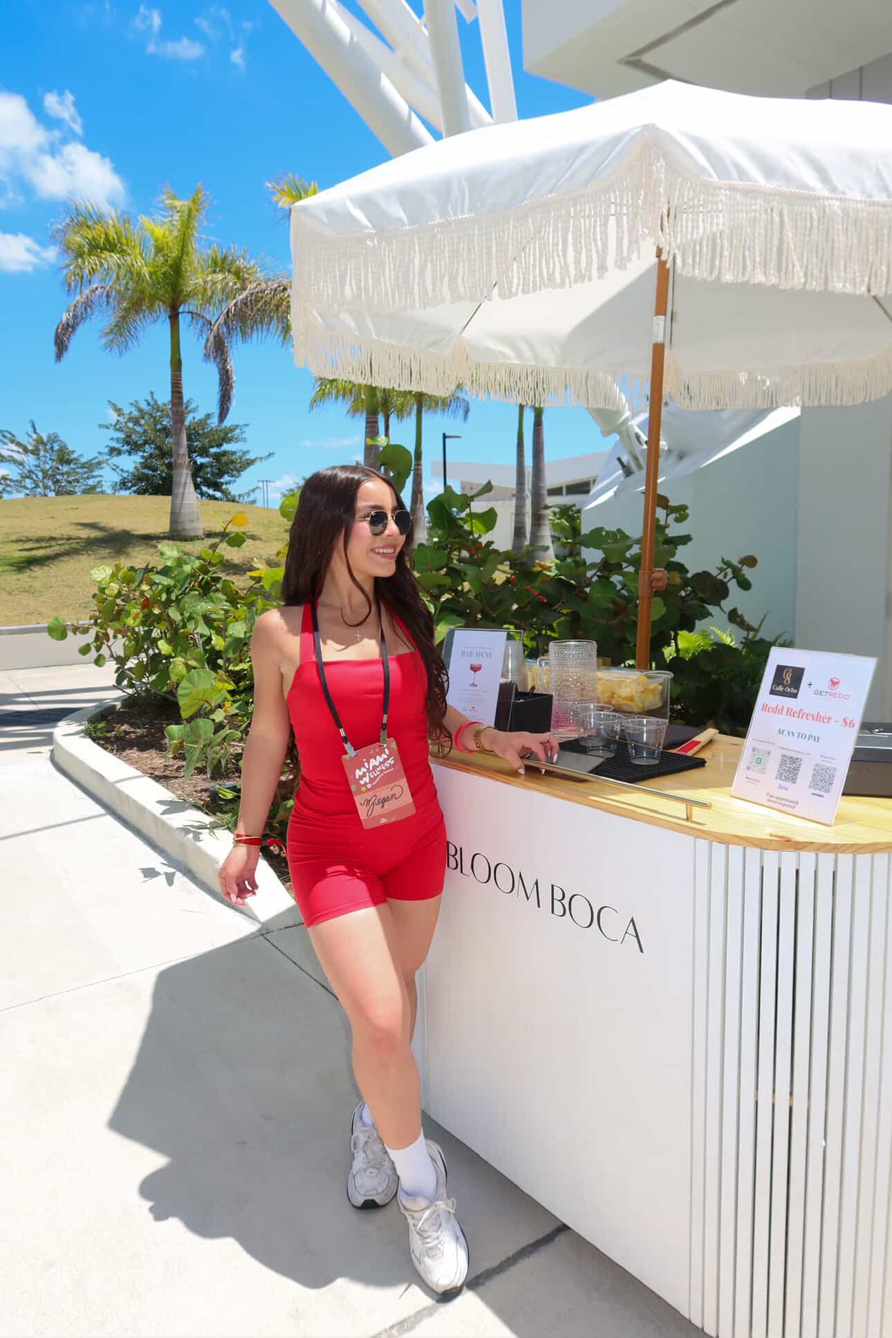 A woman wearing sunglasses, a red dress, and white sneakers standing at a white outdoor booth with a sign that reads "Bloom Boca." She is smiling and holding a small container near a sign advertising a drink called "Reald Refresher" priced at $6. There is a large white umbrella shading the booth, and in the background, there are palm trees, a grassy hill, and a partly cloudy blue sky.