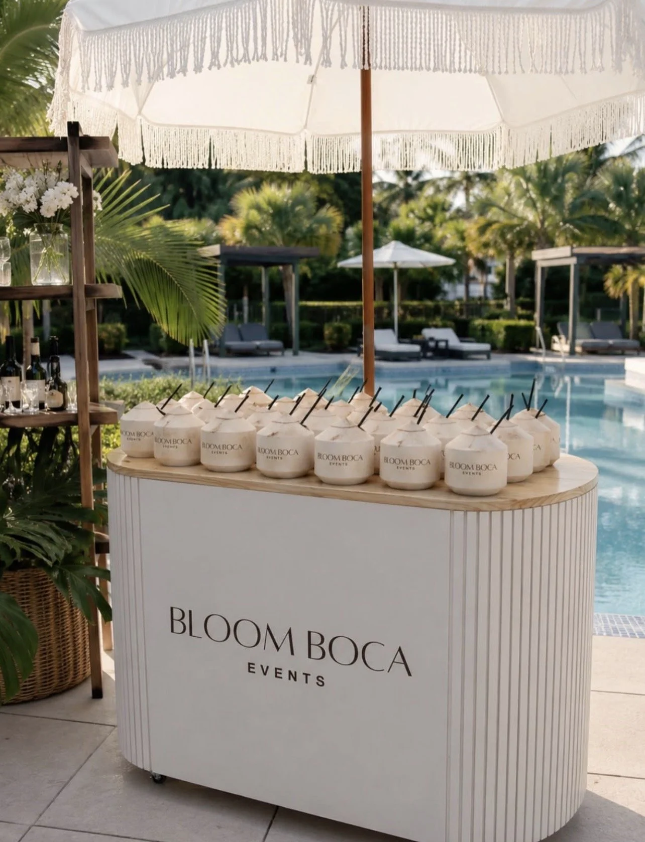 A beach club drink cart with coconuts and a large white umbrella, near a pool area with lounge chairs and lush greenery.