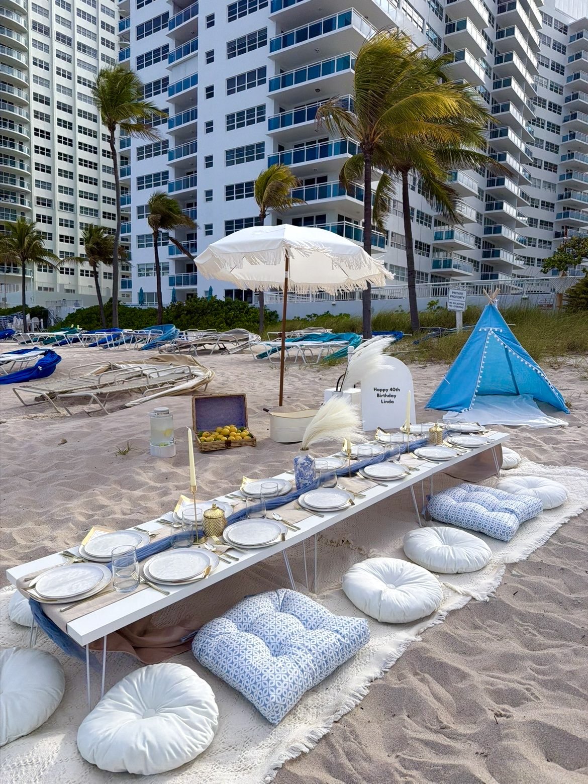 A beach setup with a low dining table, cushions, and a small tent, set on the sandy shore near tall residential buildings and palm trees.