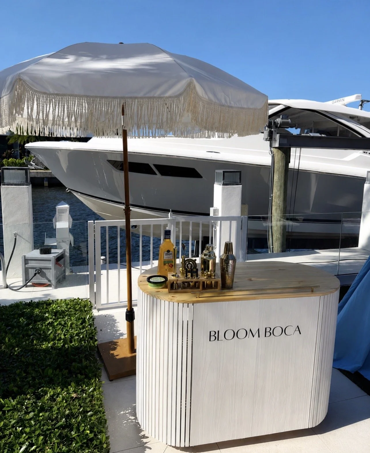 A white bar with the words 'BLOOM BOCA' is set on a dock next to a yacht. On the bar, there are bottles, a shaker, and bar tools, with a large white parasol overhead. The background shows a boat and water, under a clear blue sky.
