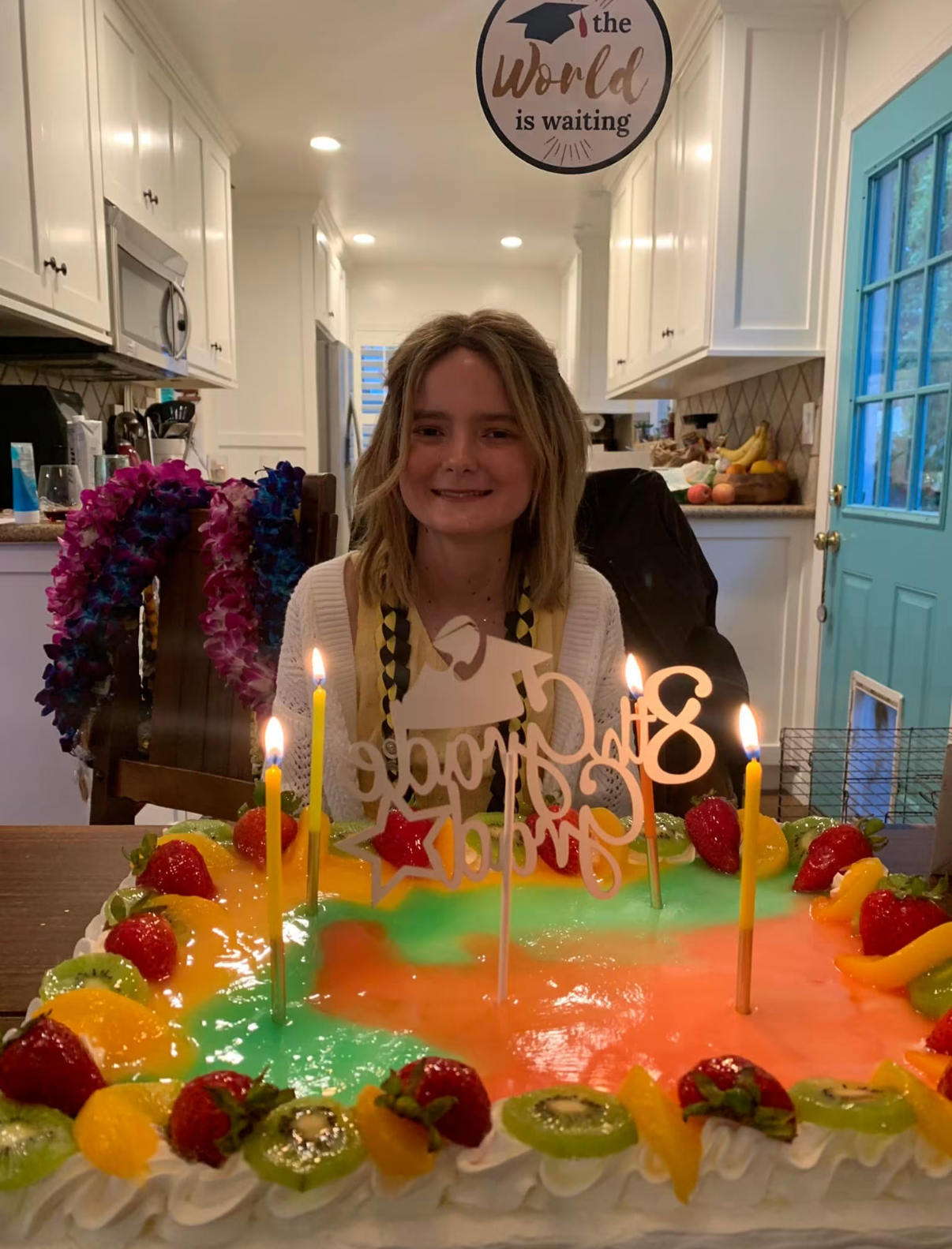 A young girl with shoulder-length blond hair sitting in front of a colorful birthday cake with lit candles, in a kitchen with white cabinets, a blue door, and a sign that reads 'the world is waiting' hanging from the ceiling.