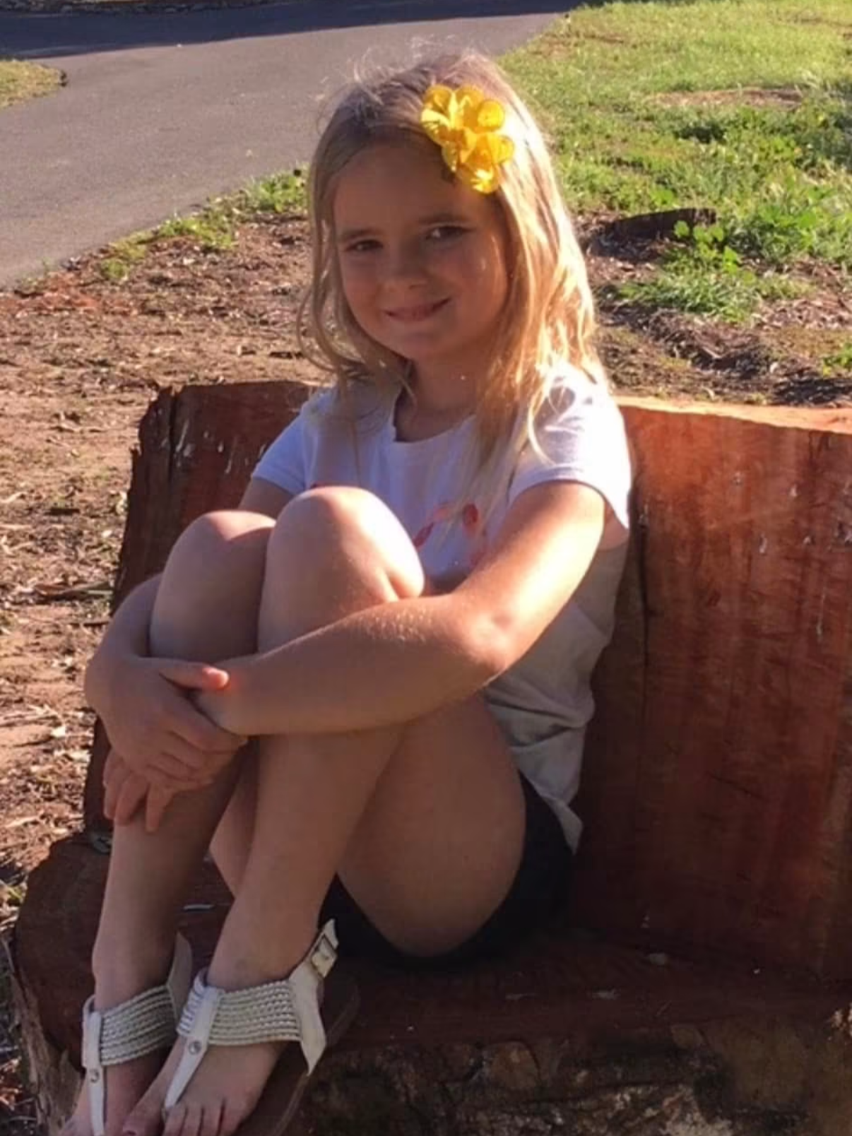 A young girl with blonde hair, wearing a yellow hair bow and a white T-shirt, sitting on a large tree trunk outdoors on a sunny day.