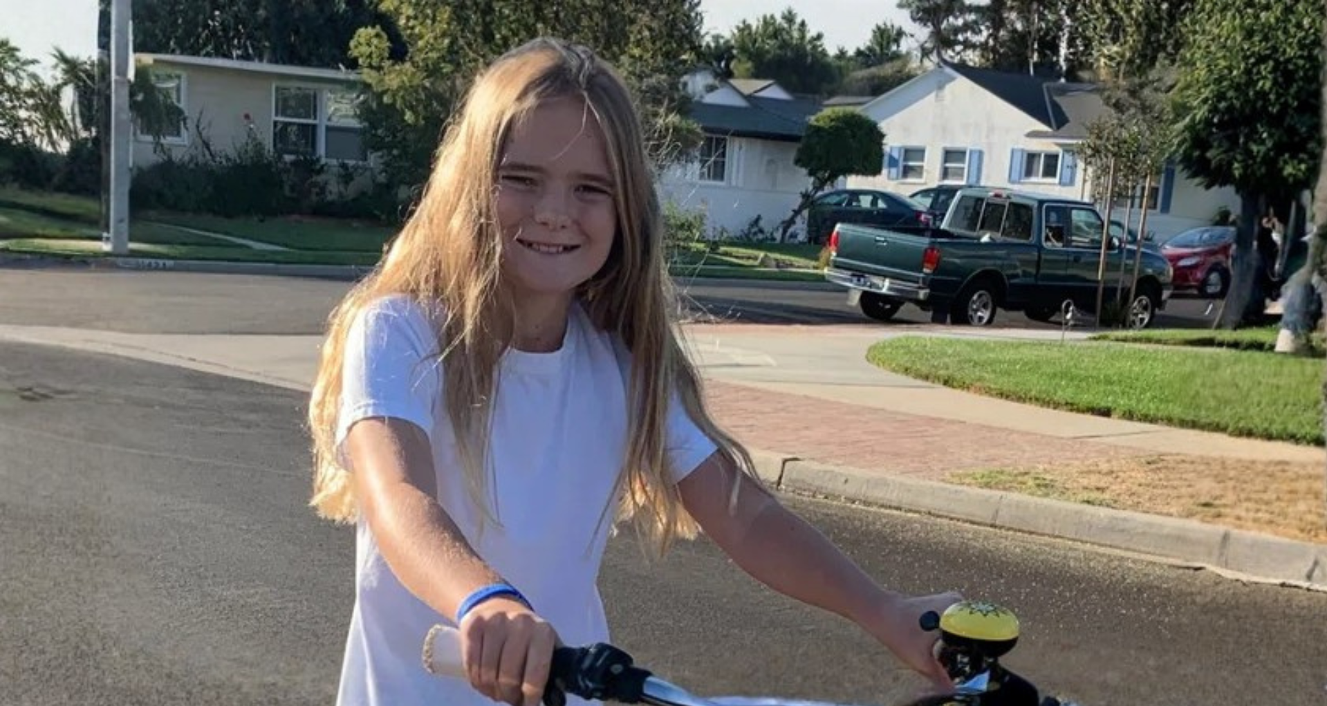 A young girl with long blonde hair riding a bicycle on a suburban street during daytime, wearing a white T-shirt, with houses, cars, and trees in the background.
