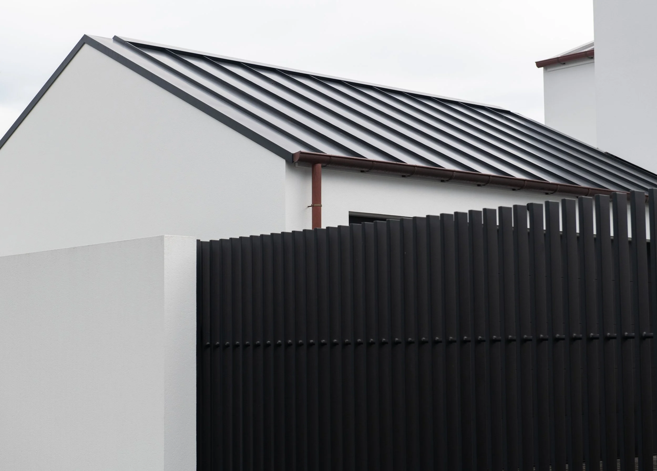 The image shows the upper part of a modern white building with a sloped roof and black metal roof panels. There is a dark brown gutter along the edge of the roof and a black wooden fence in the foreground.