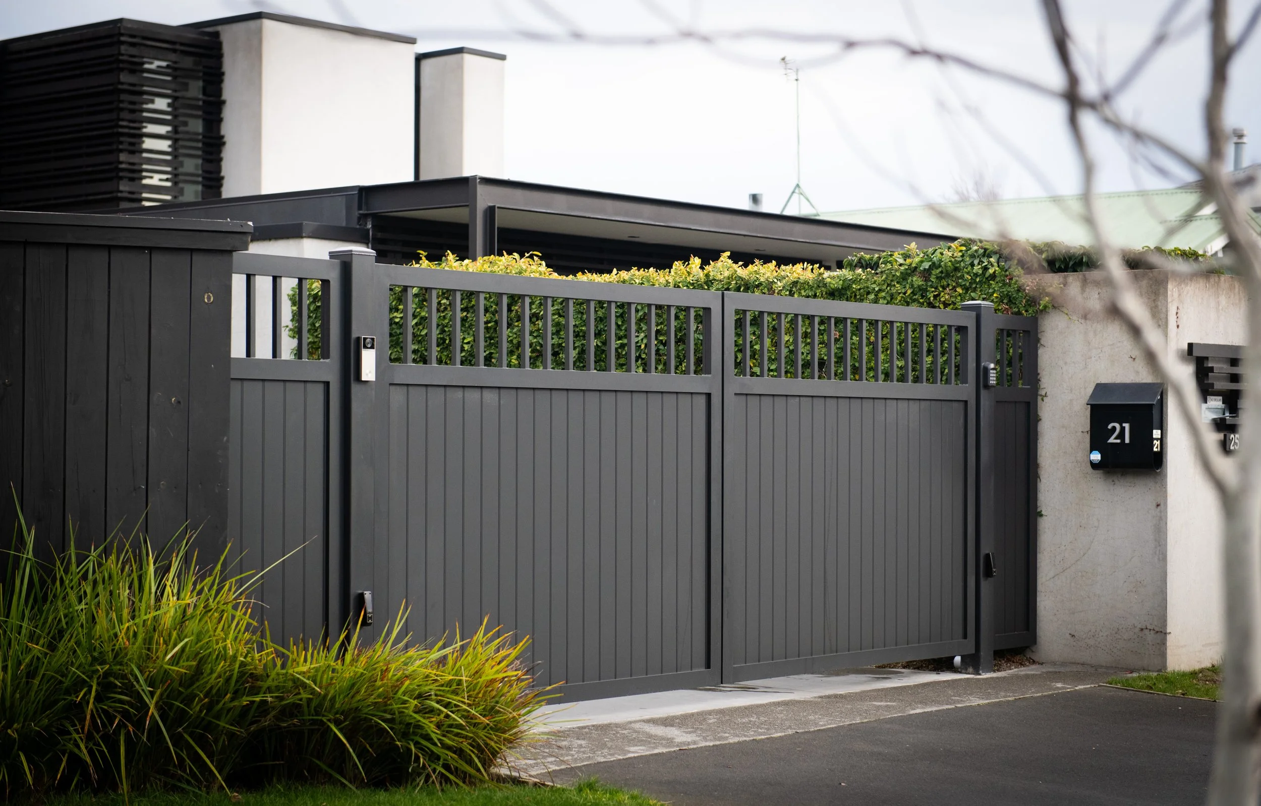 A black metal sliding gate with two mailboxes attached, situated in front of a house with a concrete wall and green foliage on top. A portion of the sidewalk and some grass with plants are visible in the foreground.