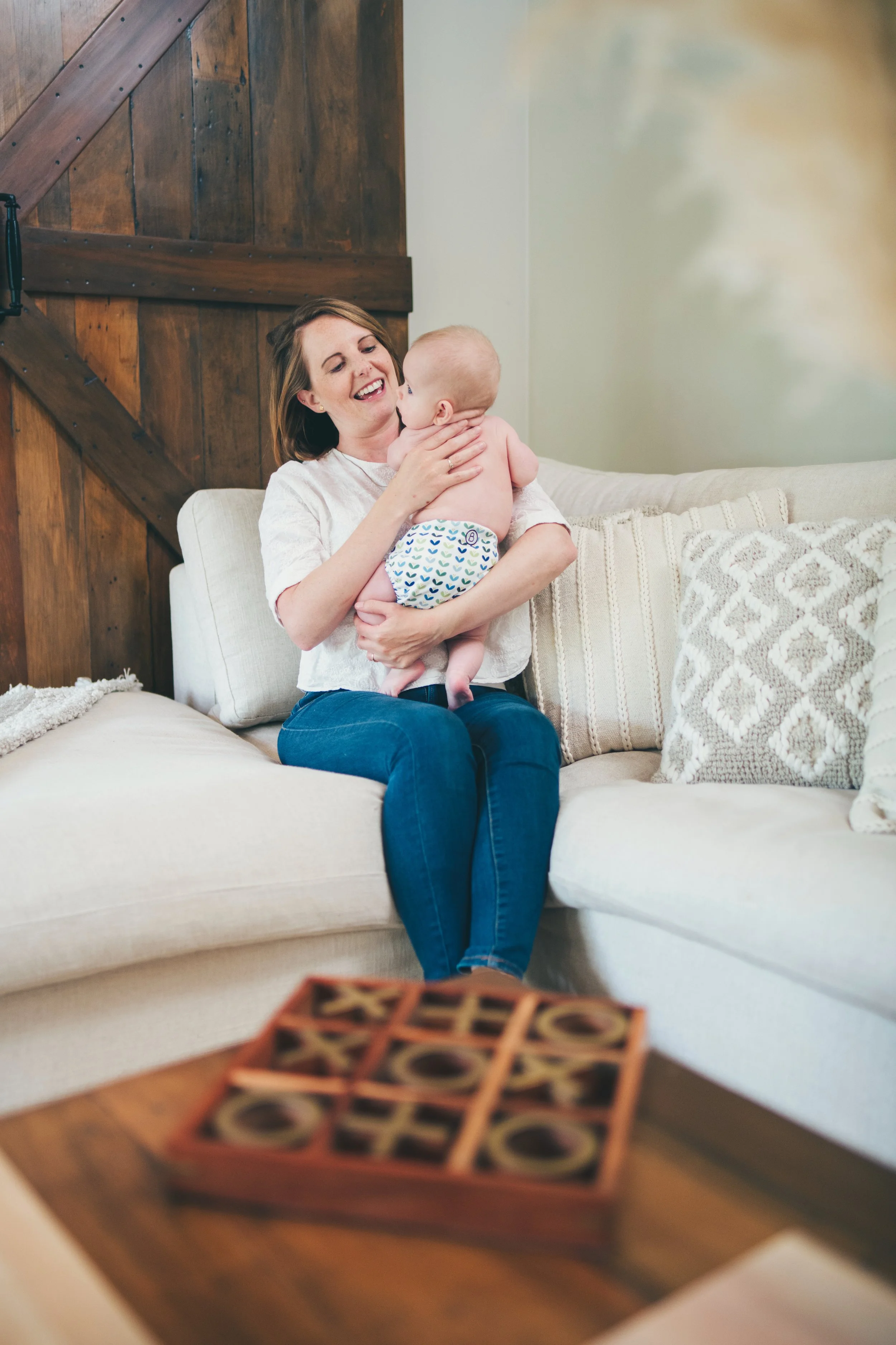 A woman with shoulder-length brown hair smiling and holding a baby dressed in a diaper, sitting on a beige sofa with textured pillows in a room with wooden wall paneling.