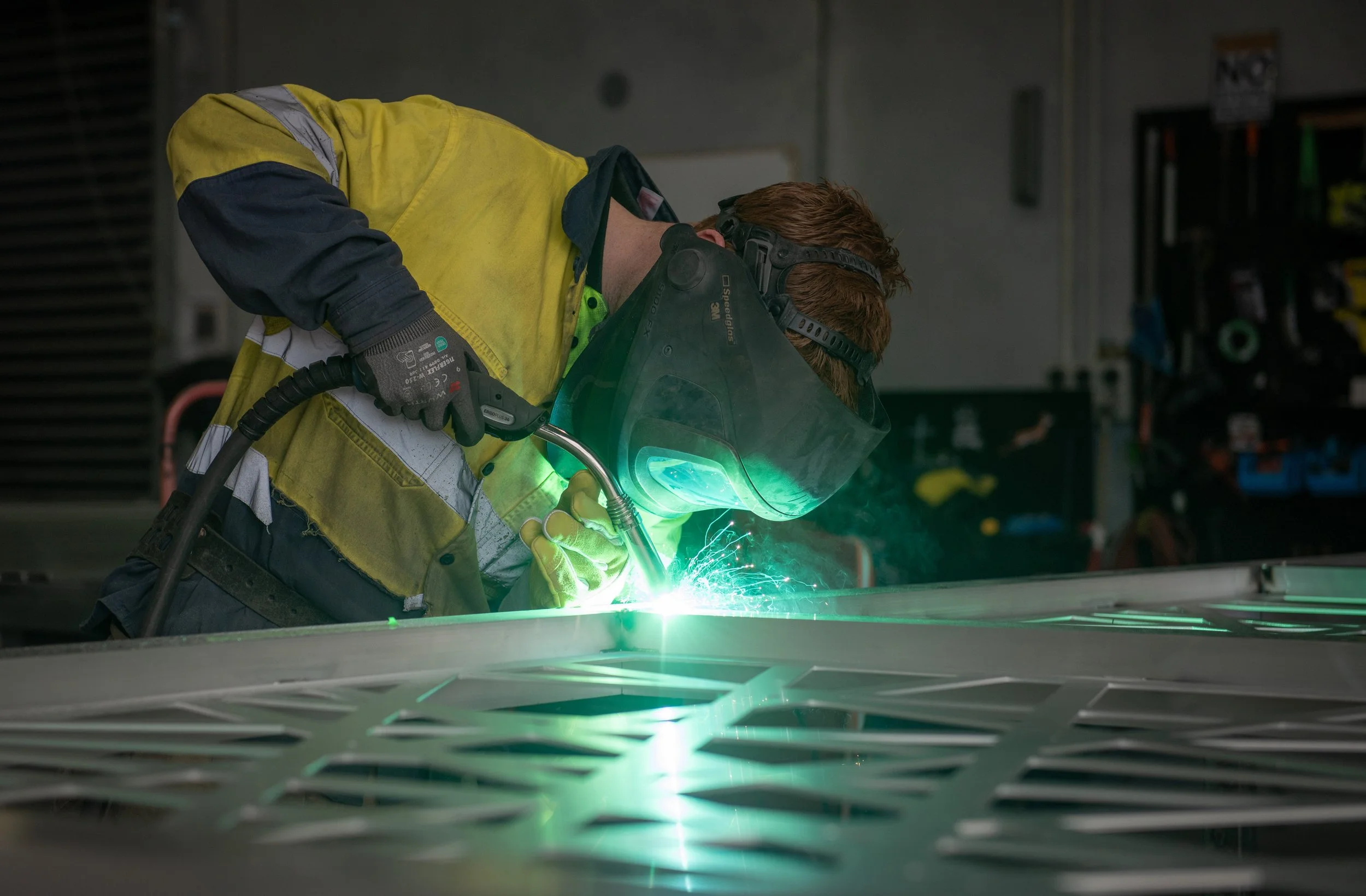 A worker wearing a yellow and black safety vest, gloves, and a welding helmet is welding metal, creating bright green sparks in an industrial setting.