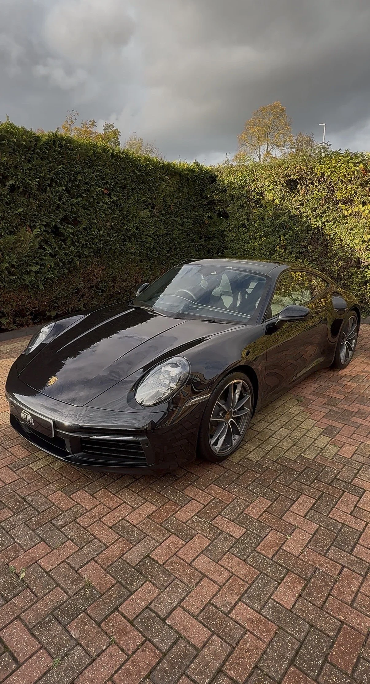 A black Porsche sports car parked on a brick driveway with green bushes and cloudy sky in the background.