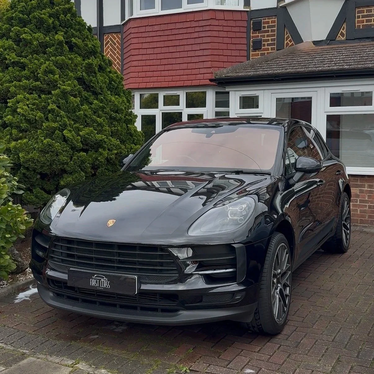 A black Porsche SUV parked on a brick driveway in front of a house with brick and white siding, next to a green bush and some plants.