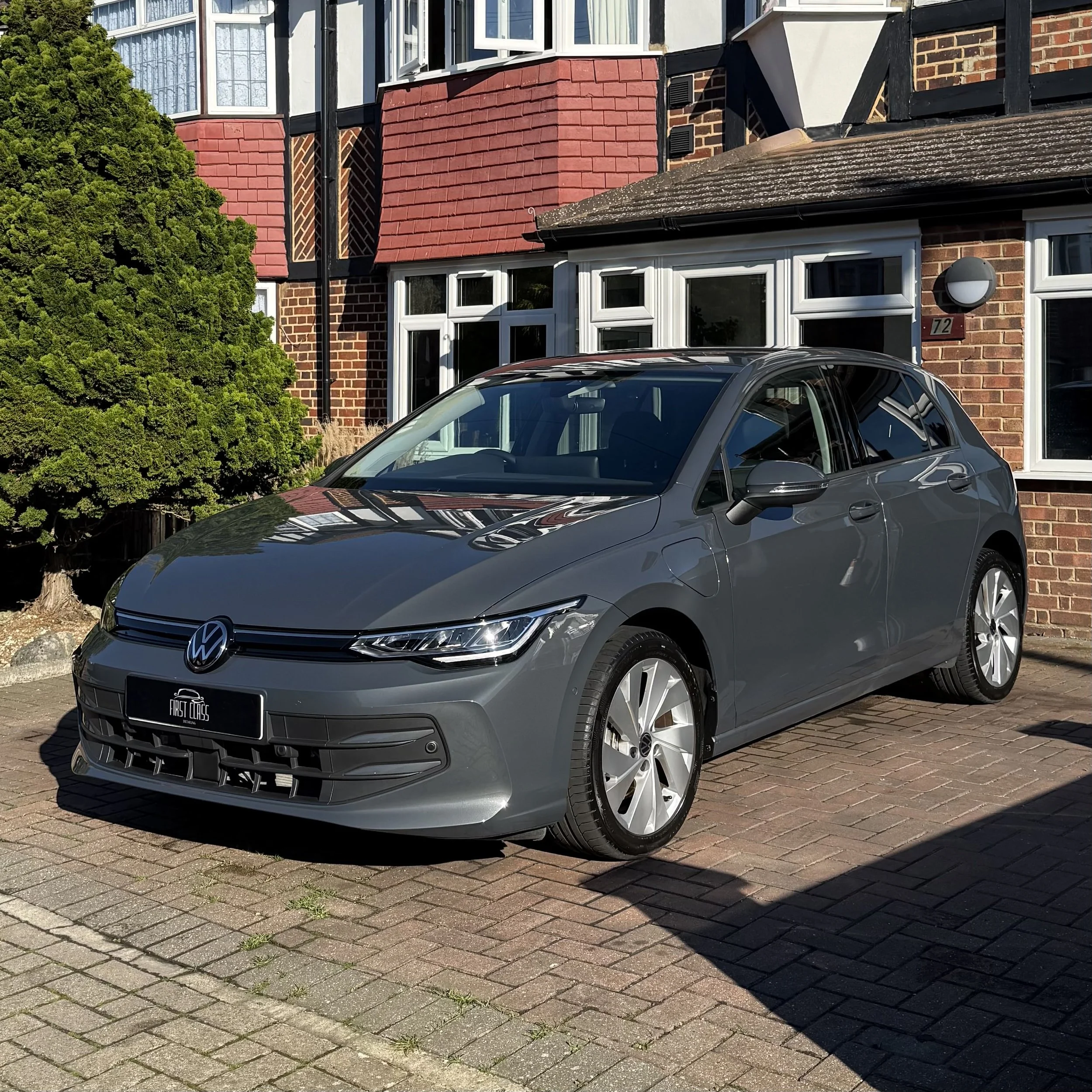A modern gray Volkswagen car parked on a brick driveway in front of a residential building with brick and half-timbered walls.