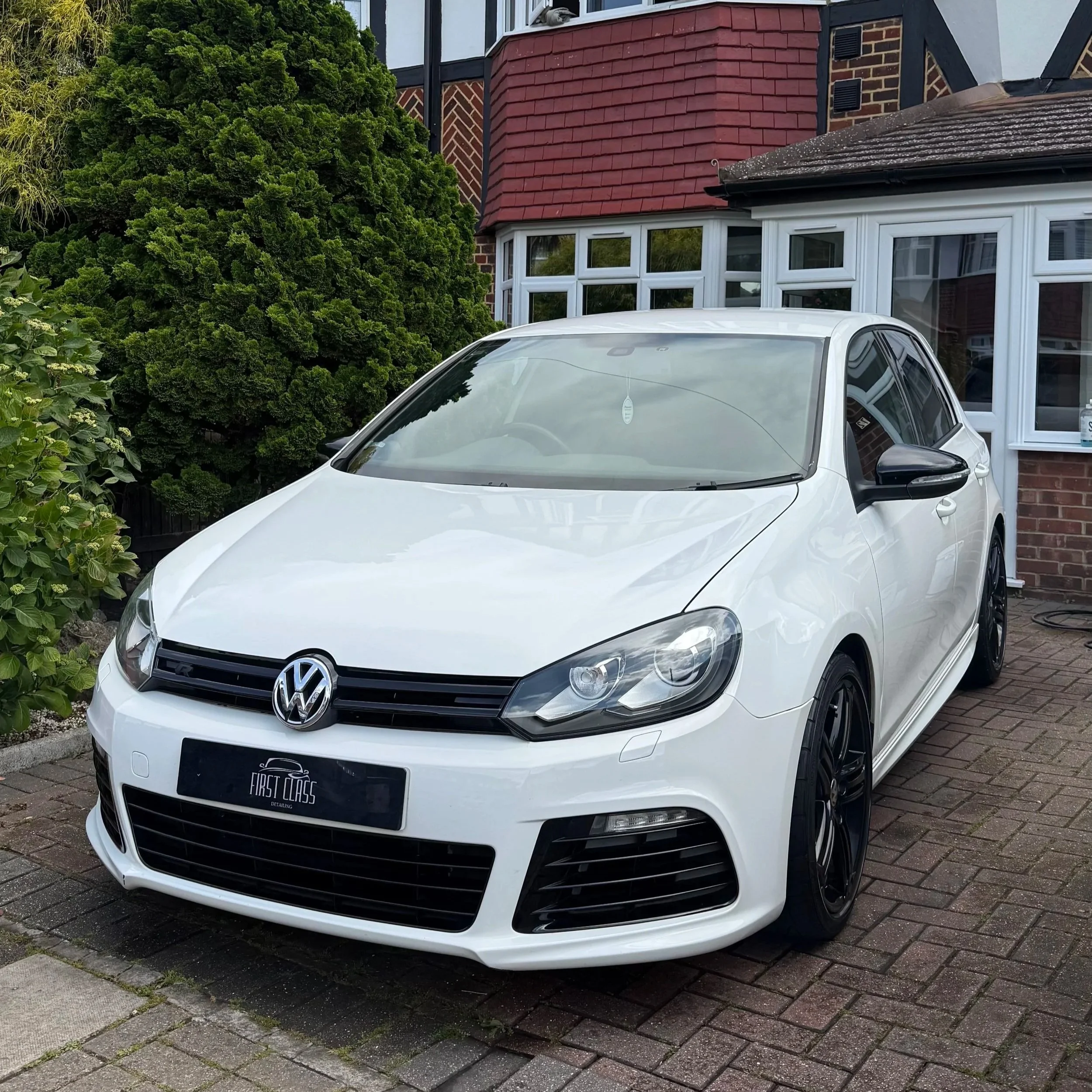 A white Volkswagen car parked in front of a house with a brick and painted exterior. The house has red shingle roofing and a bay window. Extensive green shrubbery and plants are next to the car.