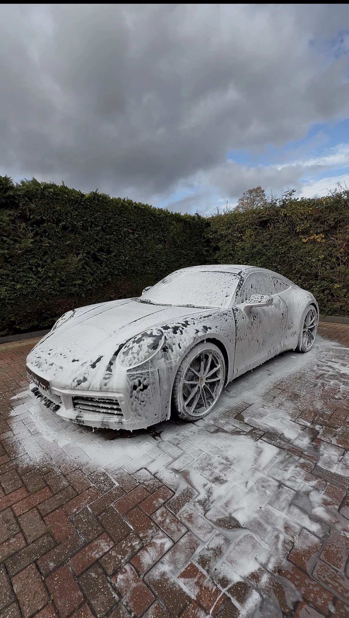A silver sports car covered in soap foam during a car wash on a paved driveway, with hedge bushes in the background and a cloudy sky overhead.