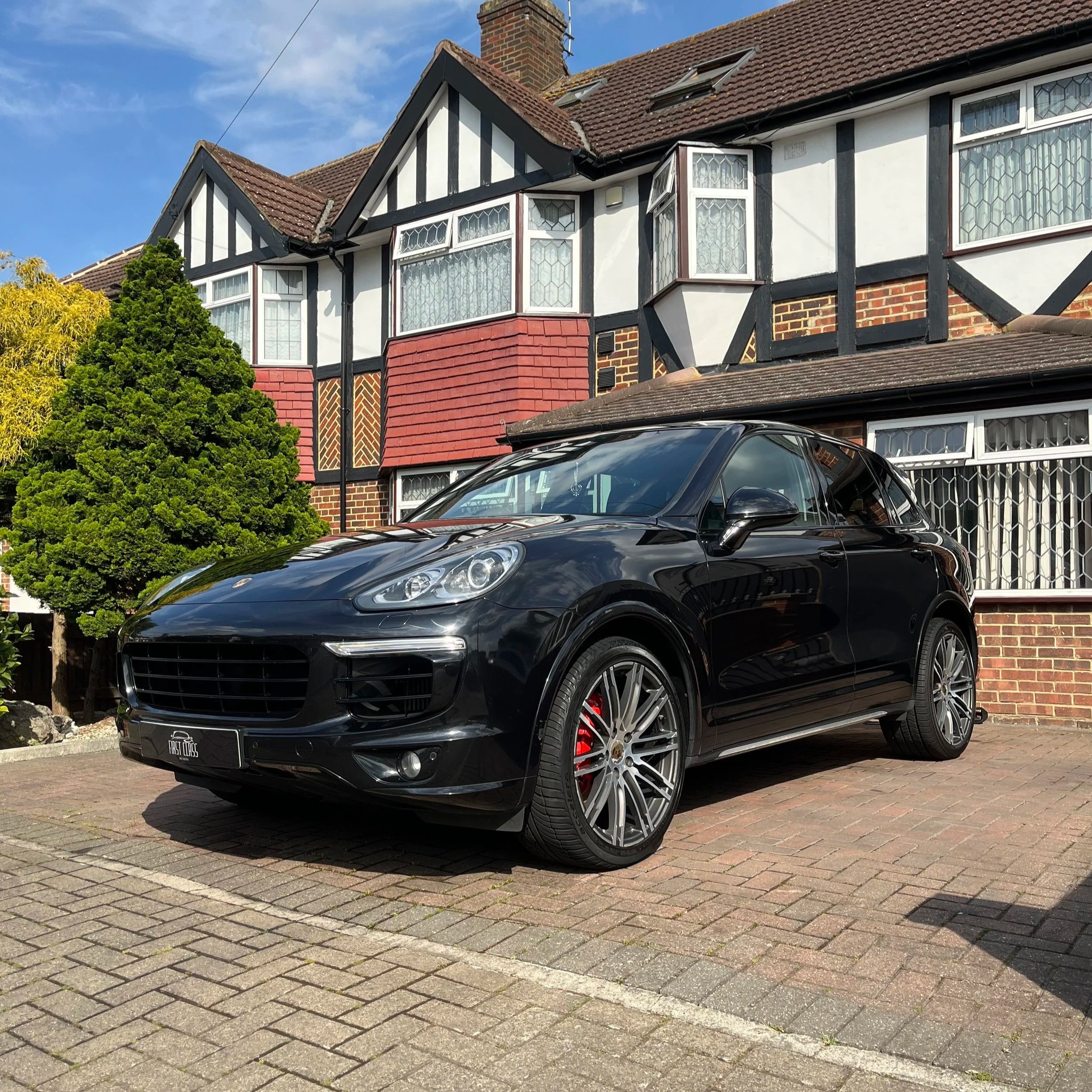 A black sports car with red brake calipers parked on a brick driveway in front of a traditional house with black-and-white timber framing and red brick walls, with a tree and a blue sky in the background.