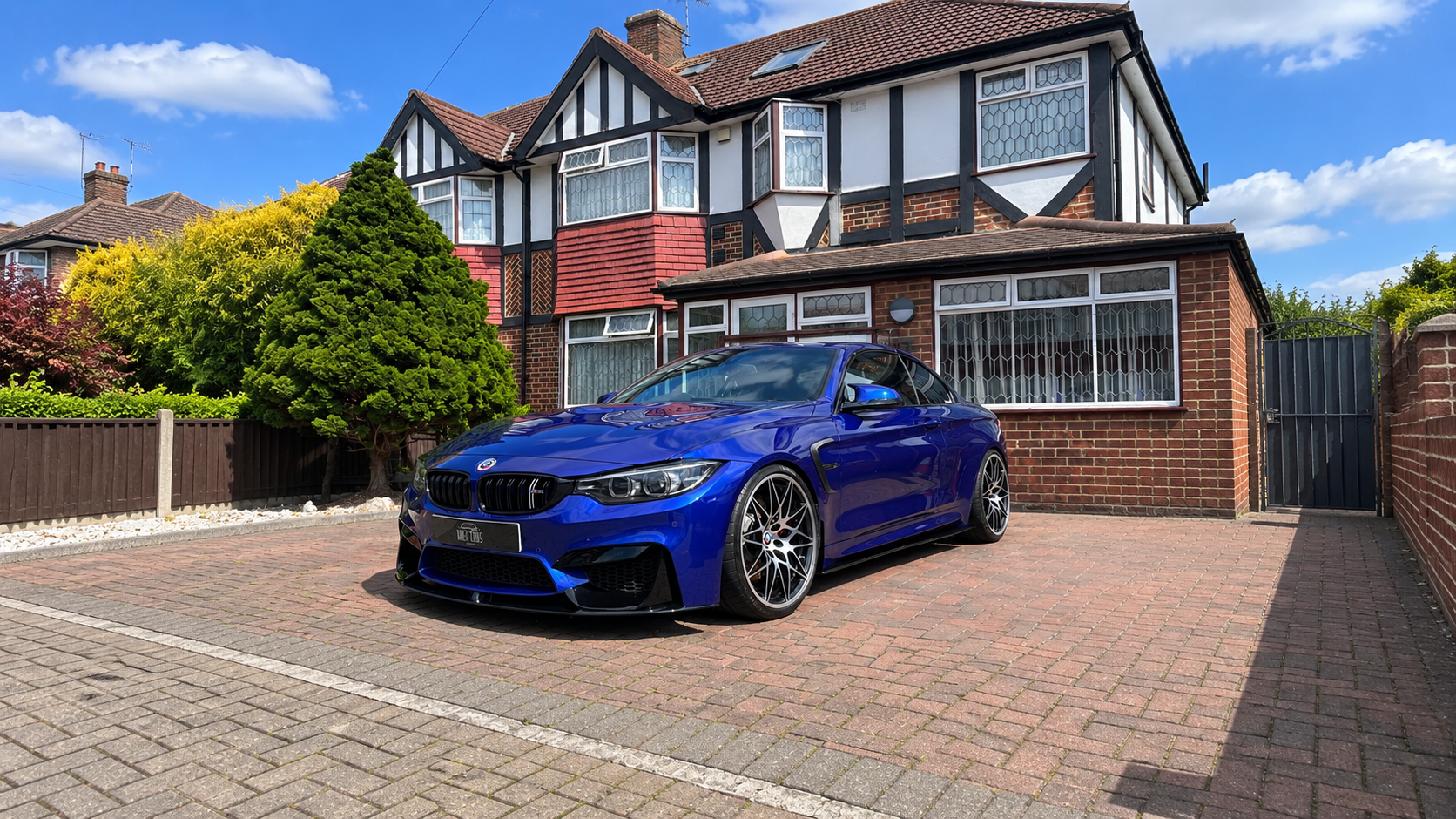 A blue sports car parked in front of a brick house with a well-maintained garden under a partly cloudy sky.