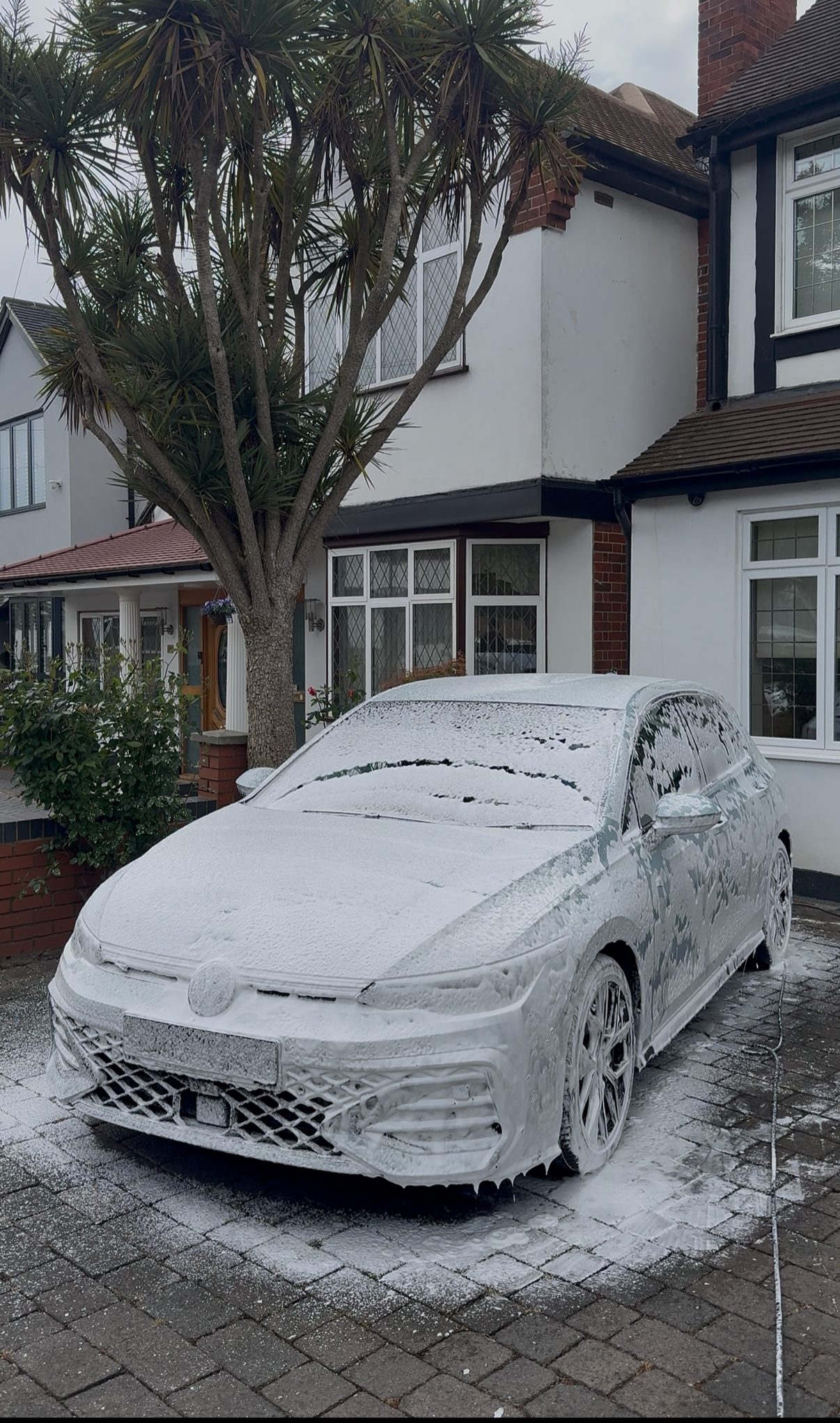 A white car covered in soap suds parked in front of a house with a tree nearby. The house has white walls, black trims, and multiple windows. The driveway is paved with bricks, and soap suds are visible on the ground.