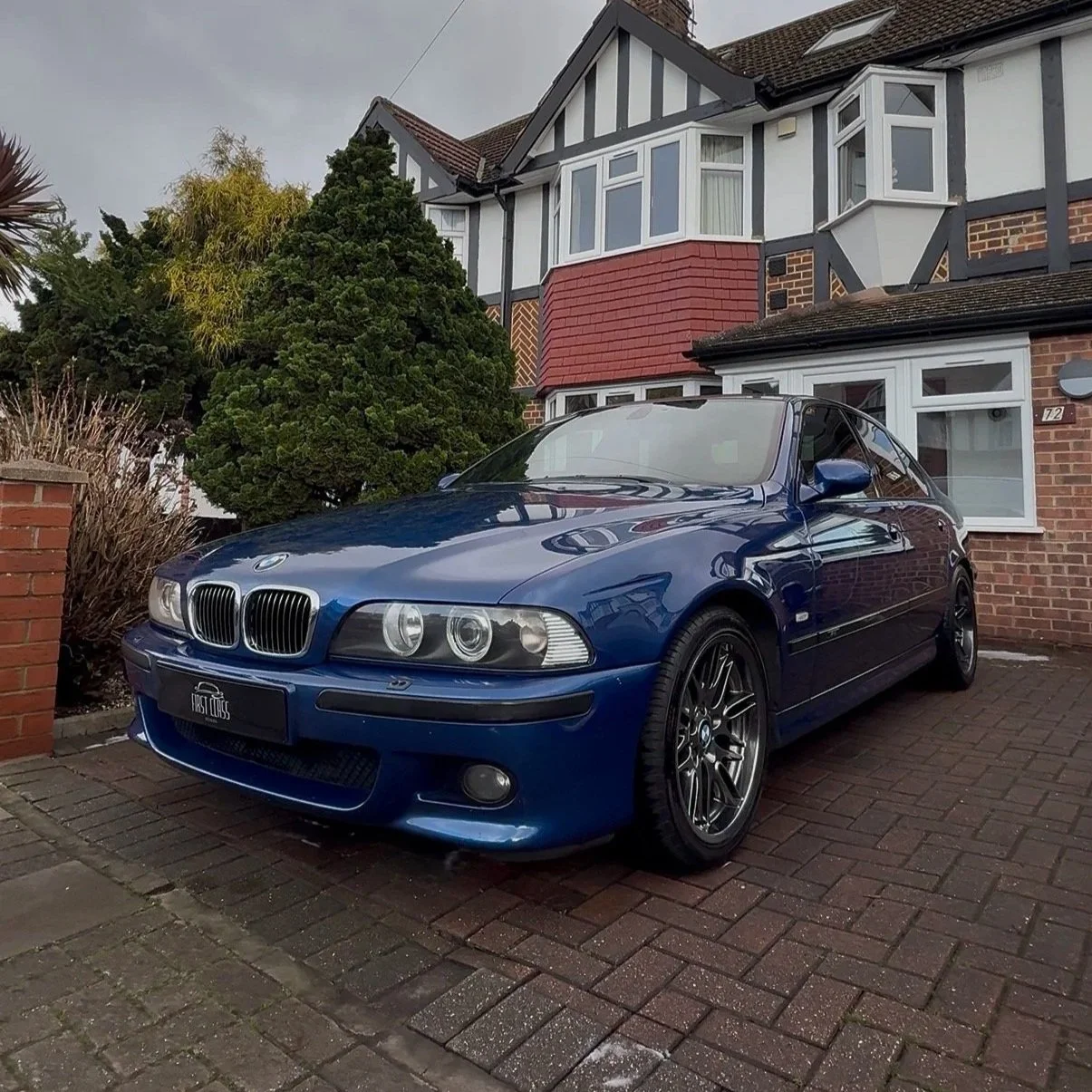 A blue BMW E46 coupe parked in front of a brick house with a garden and trees, on a brick driveway under an overcast sky.