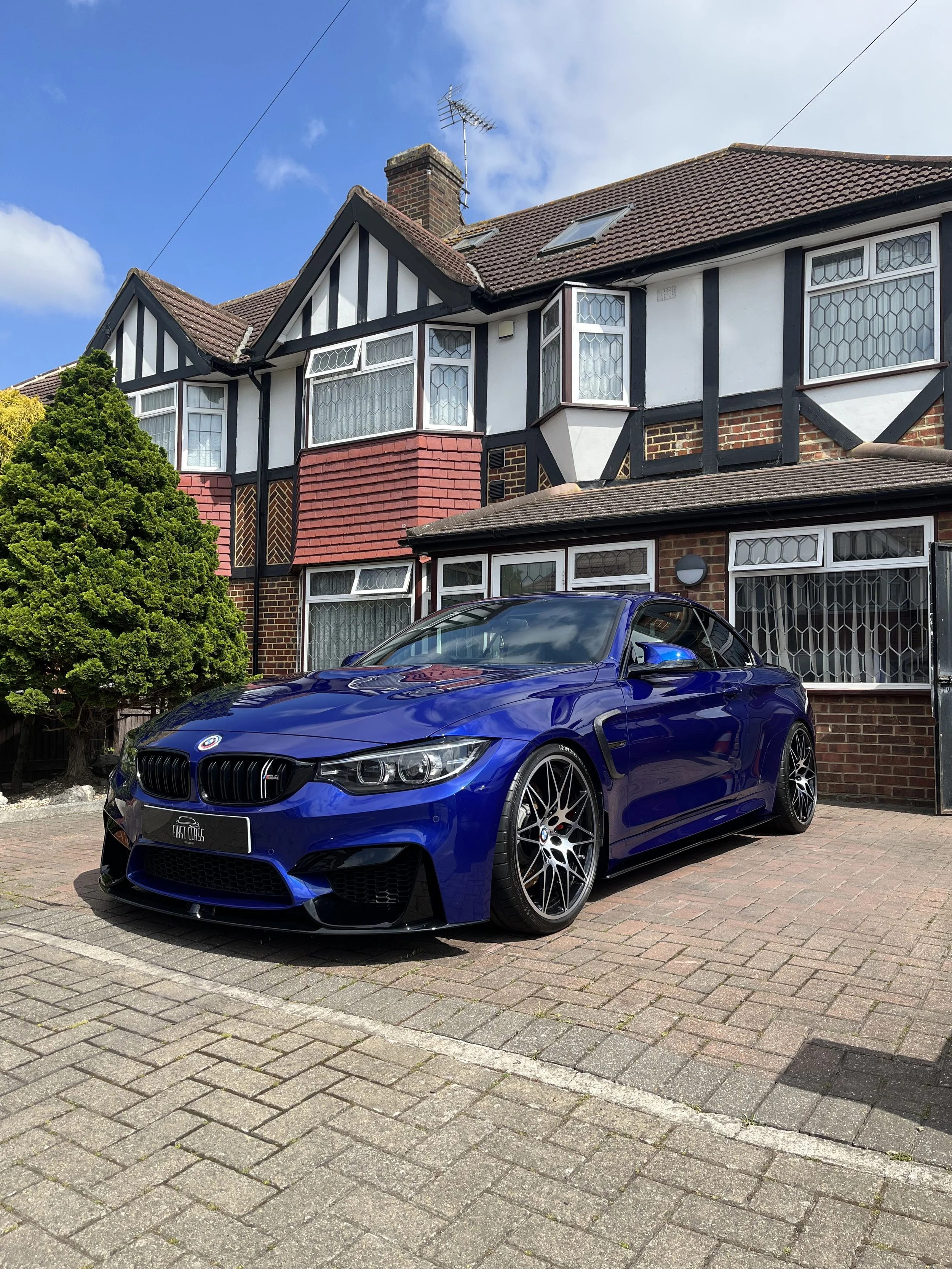 A blue BMW car parked on a brick driveway in front of a house with black and white timber framing, brickwork, and windows with leaded glass. The house has a sloped roof with skylights, a chimney, and a television antenna. A large green tree is partially visible on the left side of the image.