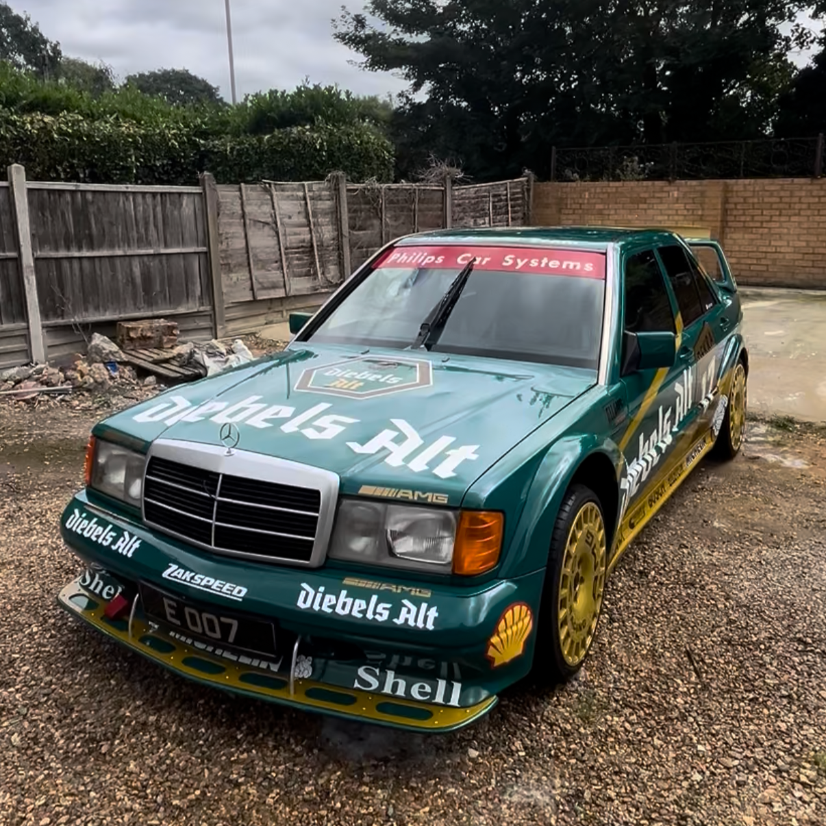 A vintage green Mercedes-Benz race car with various sponsor decals, including Shell and diebels Alt, parked on a gravel surface with a wooden fence and trees in the background.