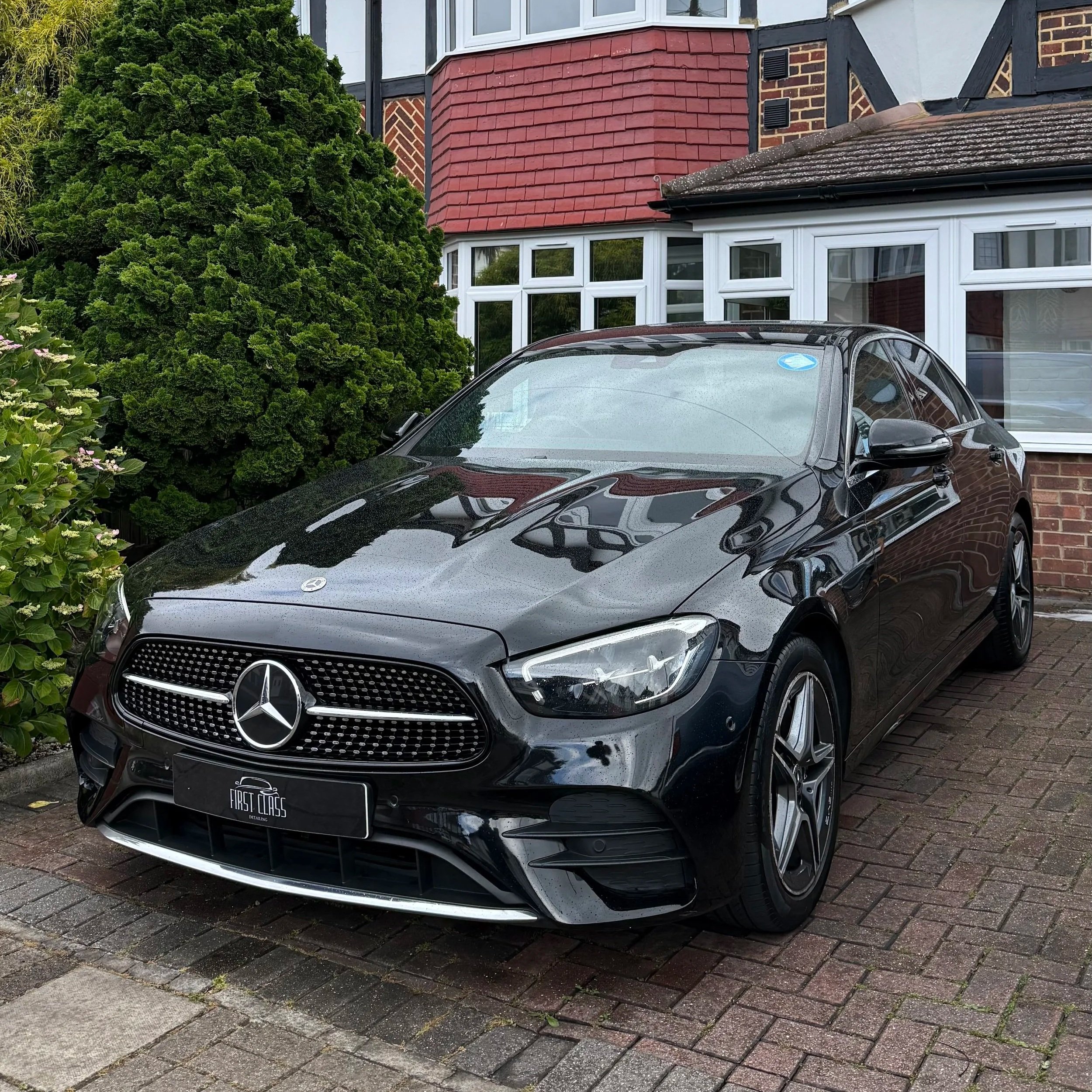 A black Mercedes-Benz parked on a driveway in front of a house with white-framed windows and red-brown shingles, surrounded by green bushes.
