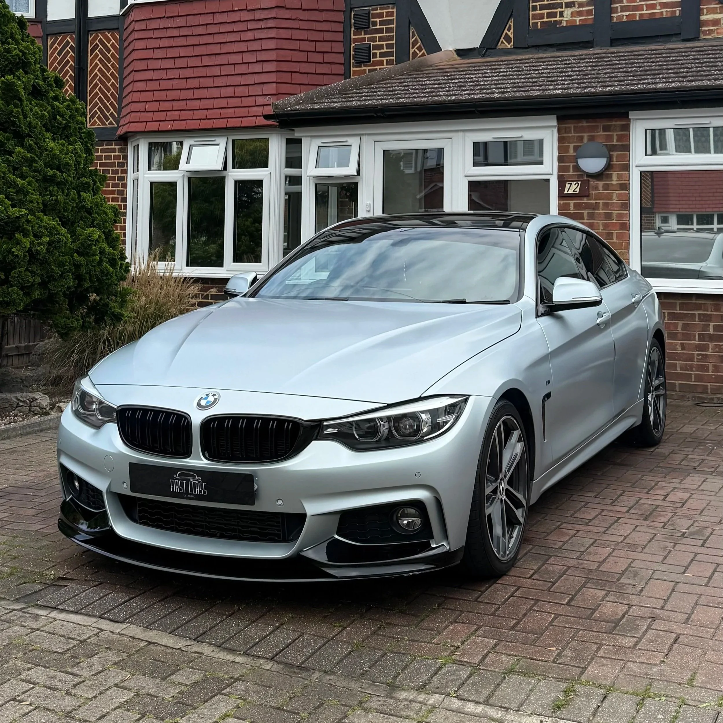 A silver BMW parked in front of a brick house with white-framed windows and a red tiled roof. The house has a house number 72 and a round external light fixture next to the window. There is greenery and landscaping on the side of the house.