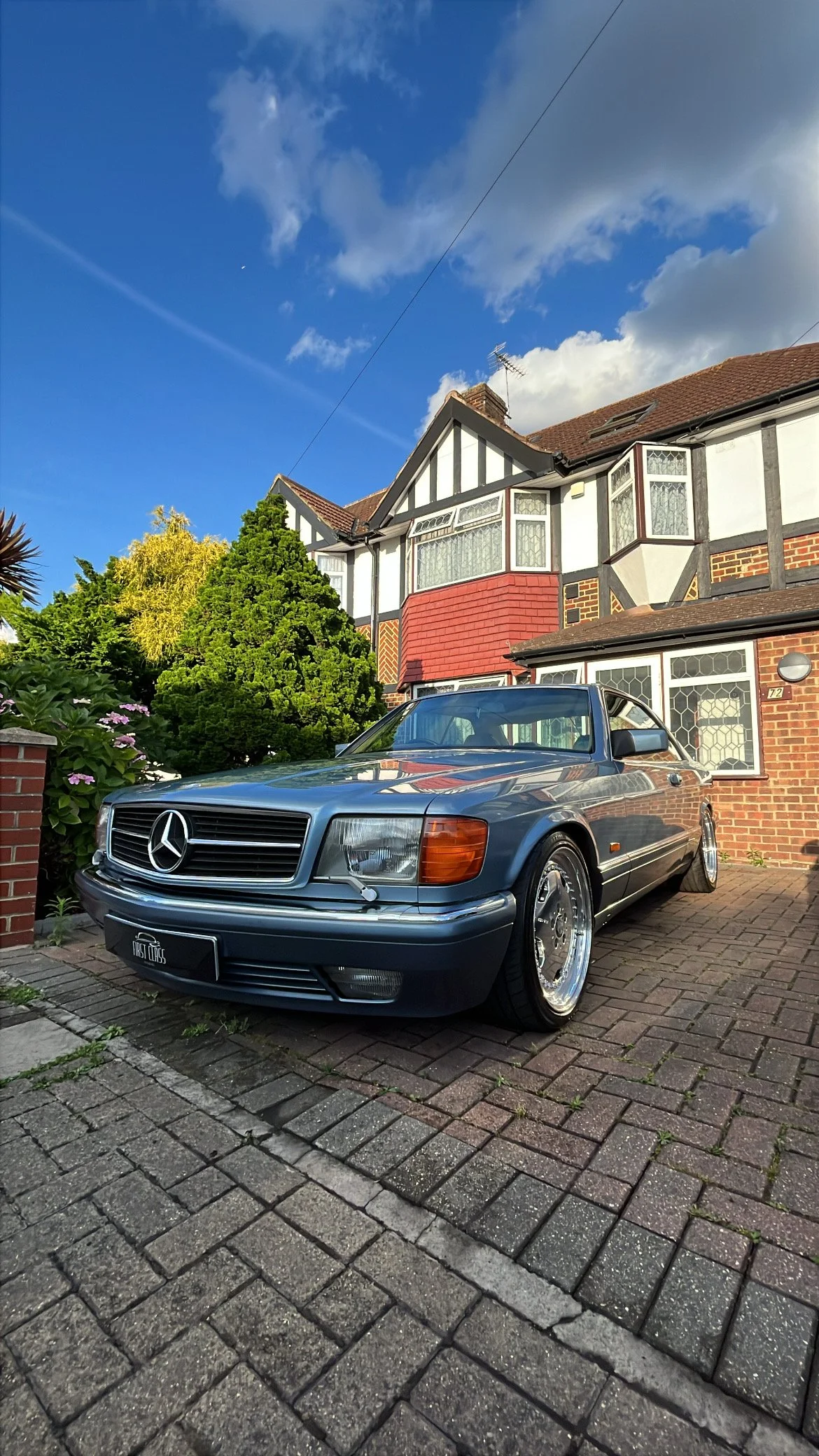 A classic Mercedes-Benz car parked in front of a brick house with white and black half-timbered details. There are green trees and flowers on the side, and a blue sky with clouds above.