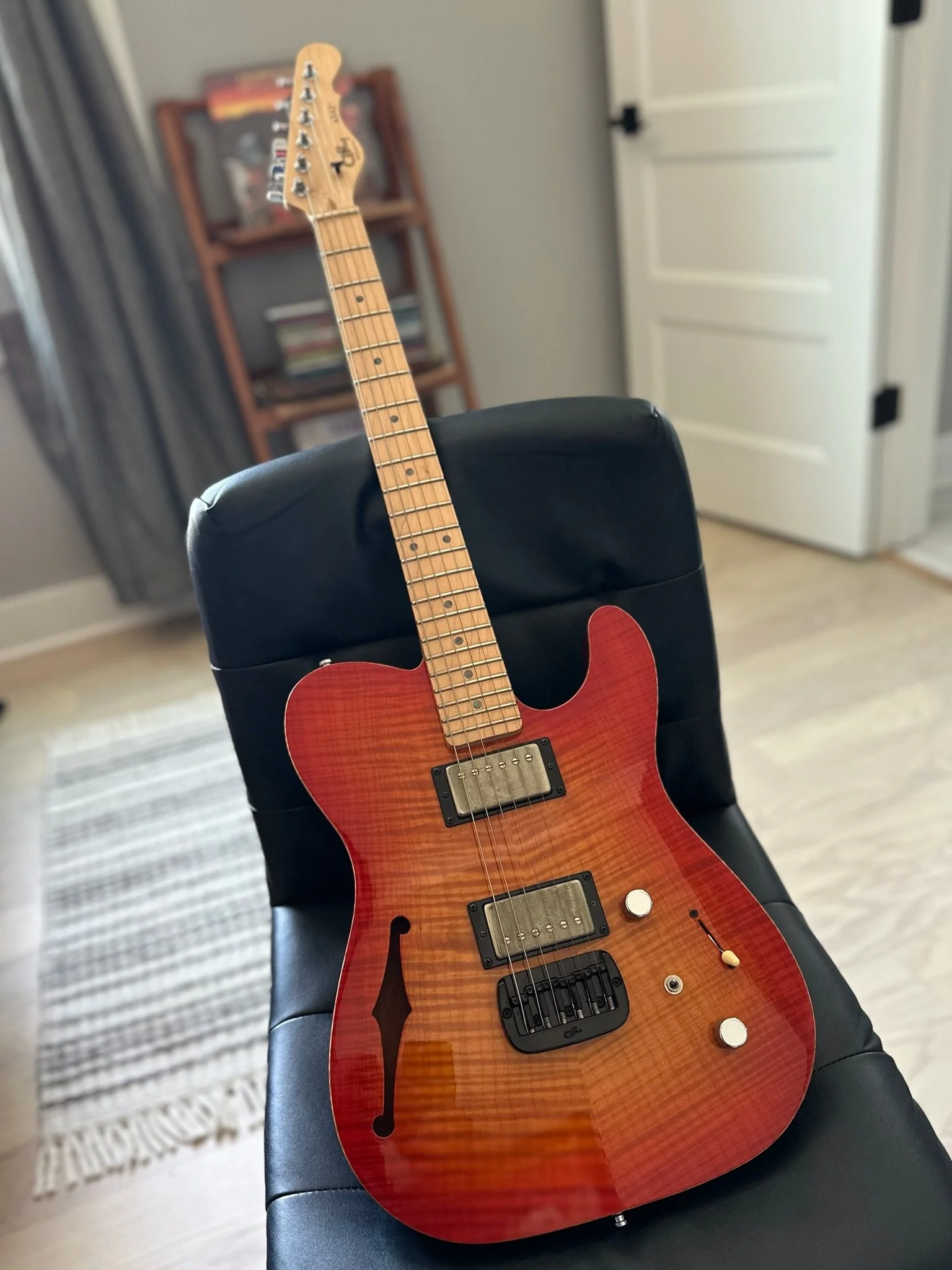 Red electric guitar resting on a black chair in a room with a bookshelf and white door in the background.