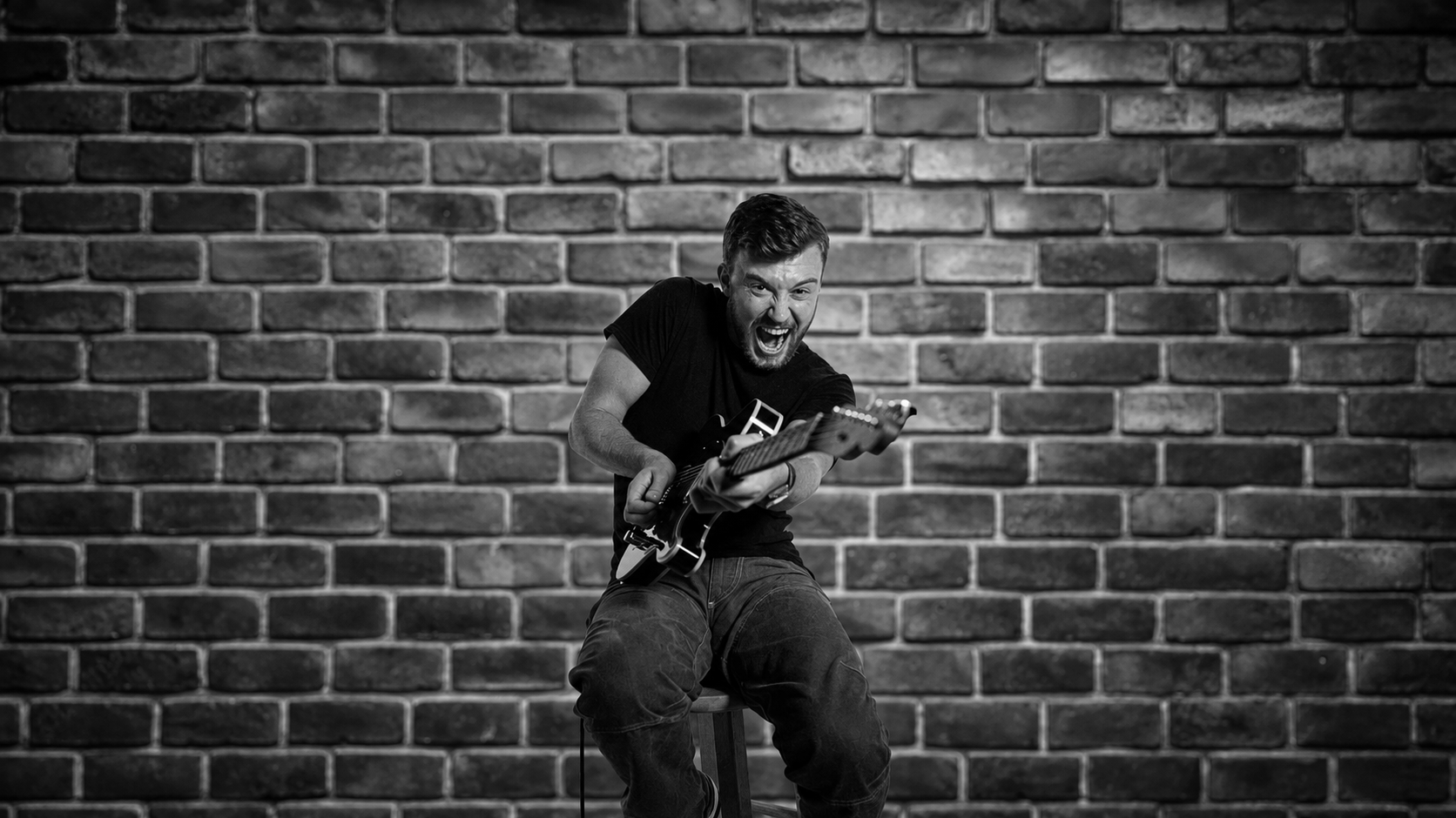 A young man with a beard in a black t-shirt and distressed jeans playing an electric guitar enthusiastically while sitting on a stool against a brick wall, in black and white.