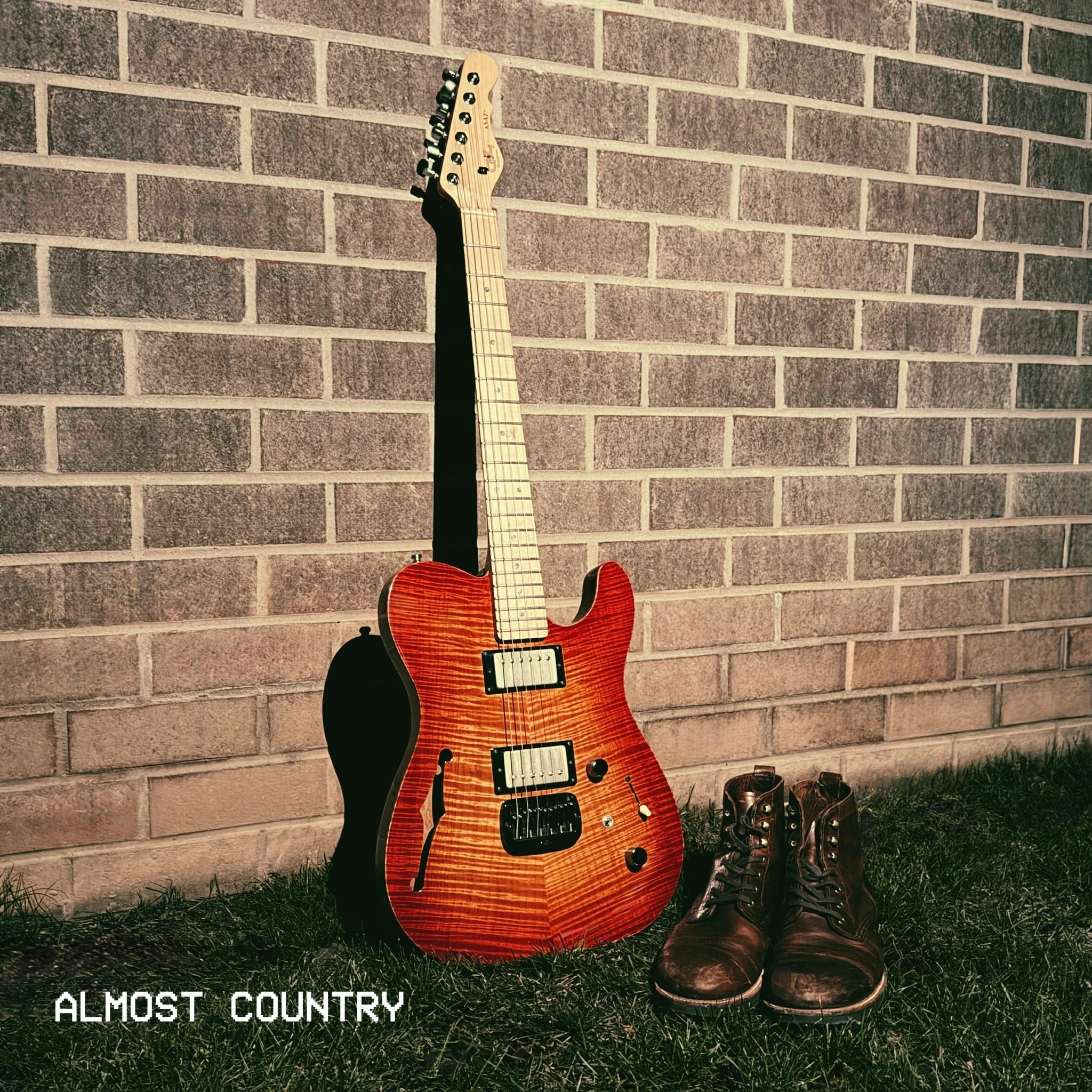 A red electric guitar leaning against a brick wall with a strap attached, and a pair of brown boots on the grass nearby.