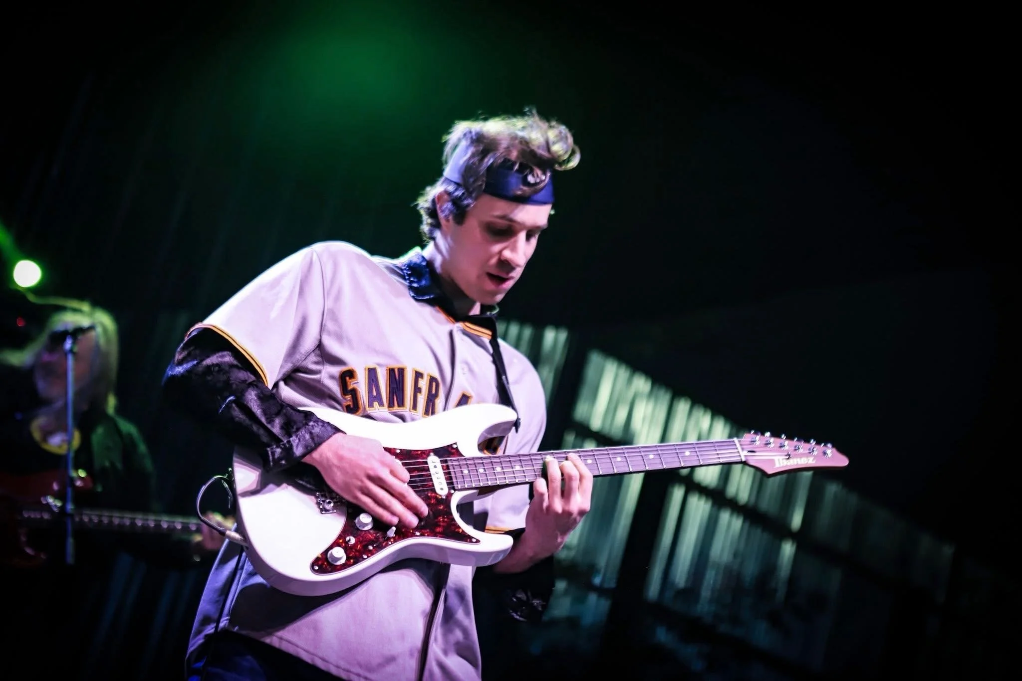 A person dressed in a San Francisco Giants baseball jersey playing an electric guitar on stage under green lighting.