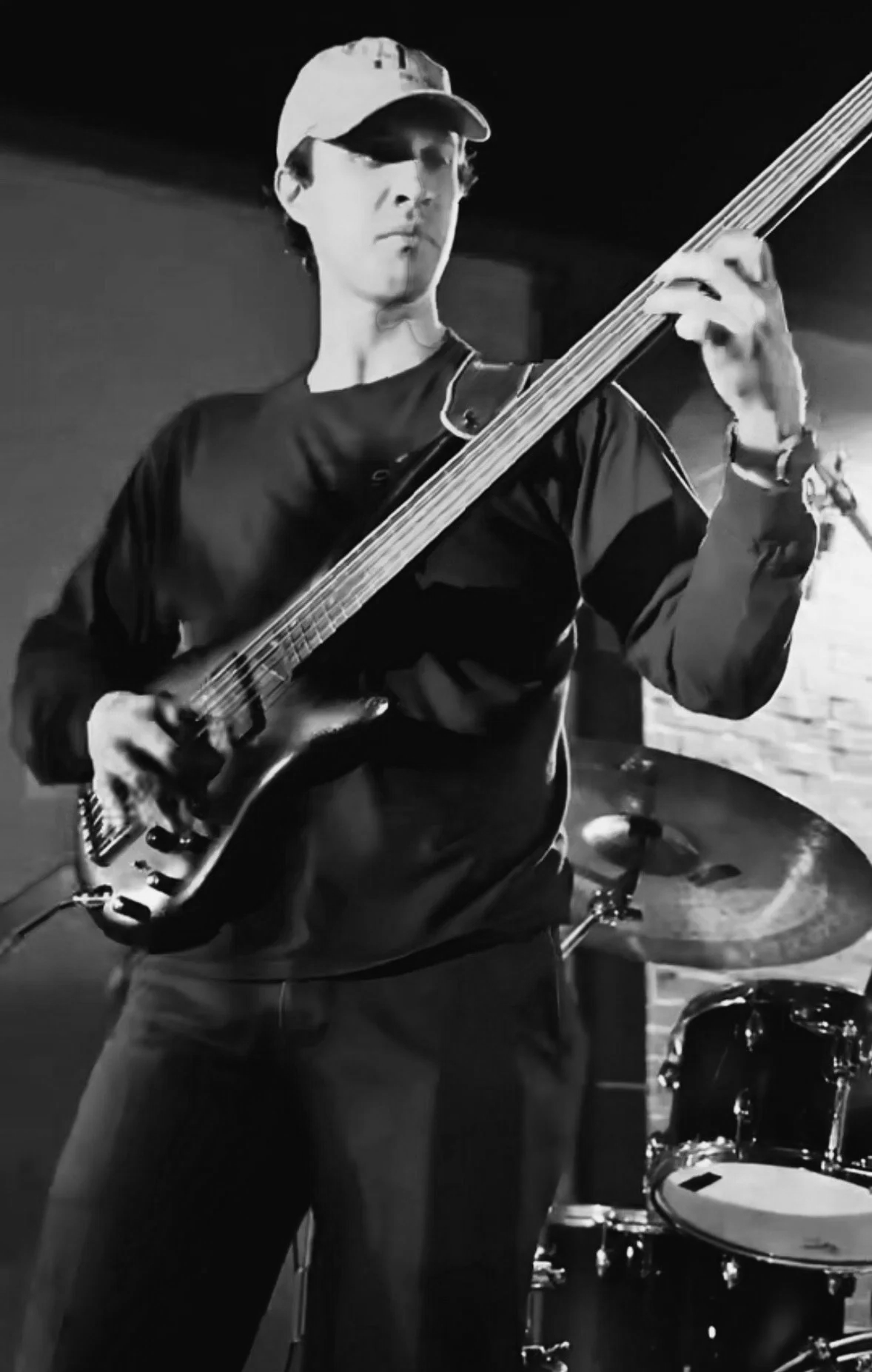 A person wearing a cap and glasses playing an electric bass guitar in a dimly lit room with a drum set in the background, black and white photo.  Nashville Guitar Lessons Zoom Guitar Lessons Virtual Lessons In Person Guitar Lessons Session Work Audit