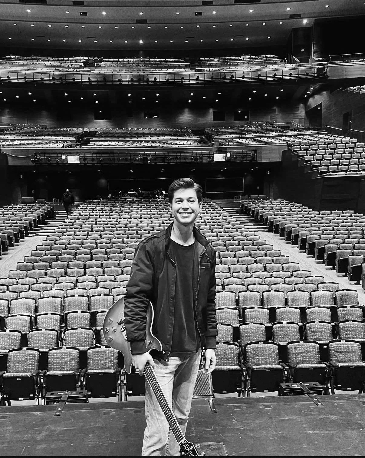 A young man standing on a stage holding a guitar in an empty concert hall with multiple rows of seats.  Nashville Guitar Lessons Zoom Guitar Lessons Virtual Lessons In Person Guitar Lessons Session Work Audition Prep
