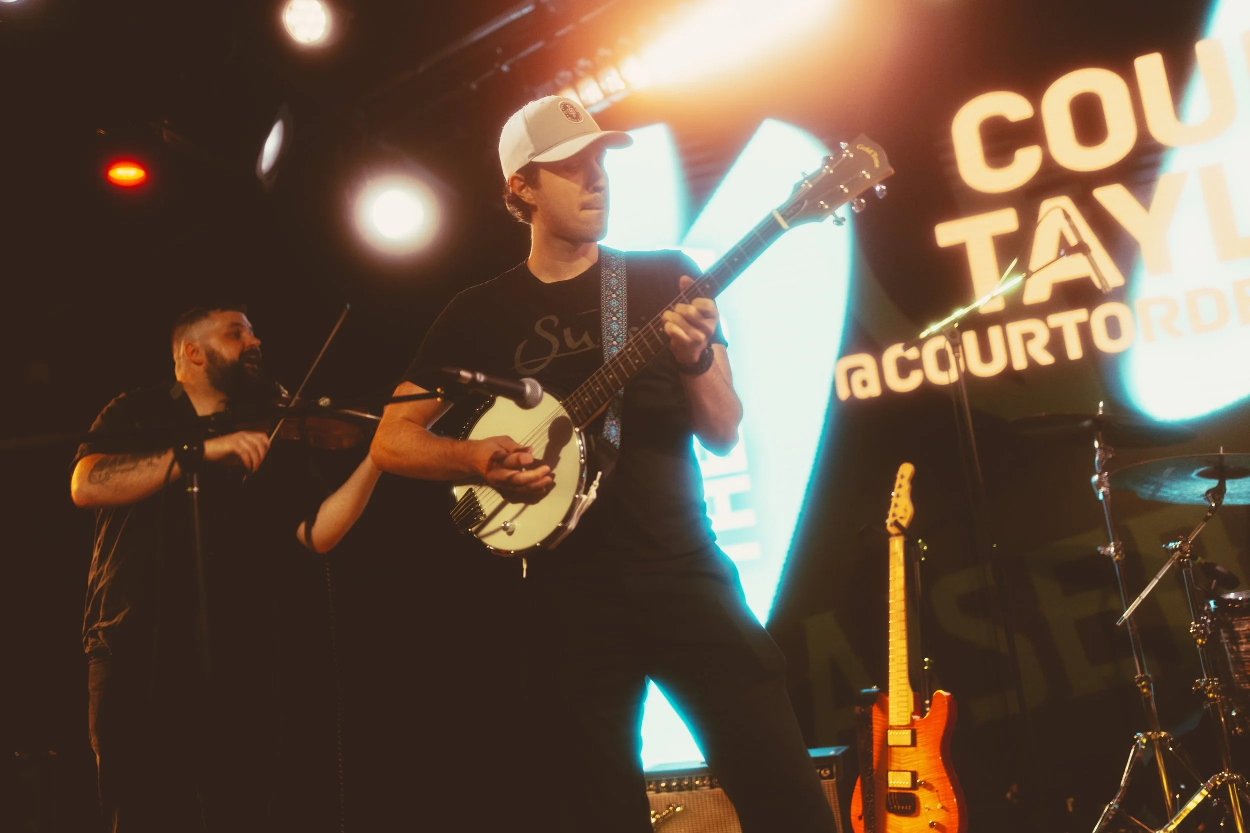 A musician playing an acoustic guitar on stage under colorful lights, with a man in a dark shirt visible in the background and a neon sign partially visible.