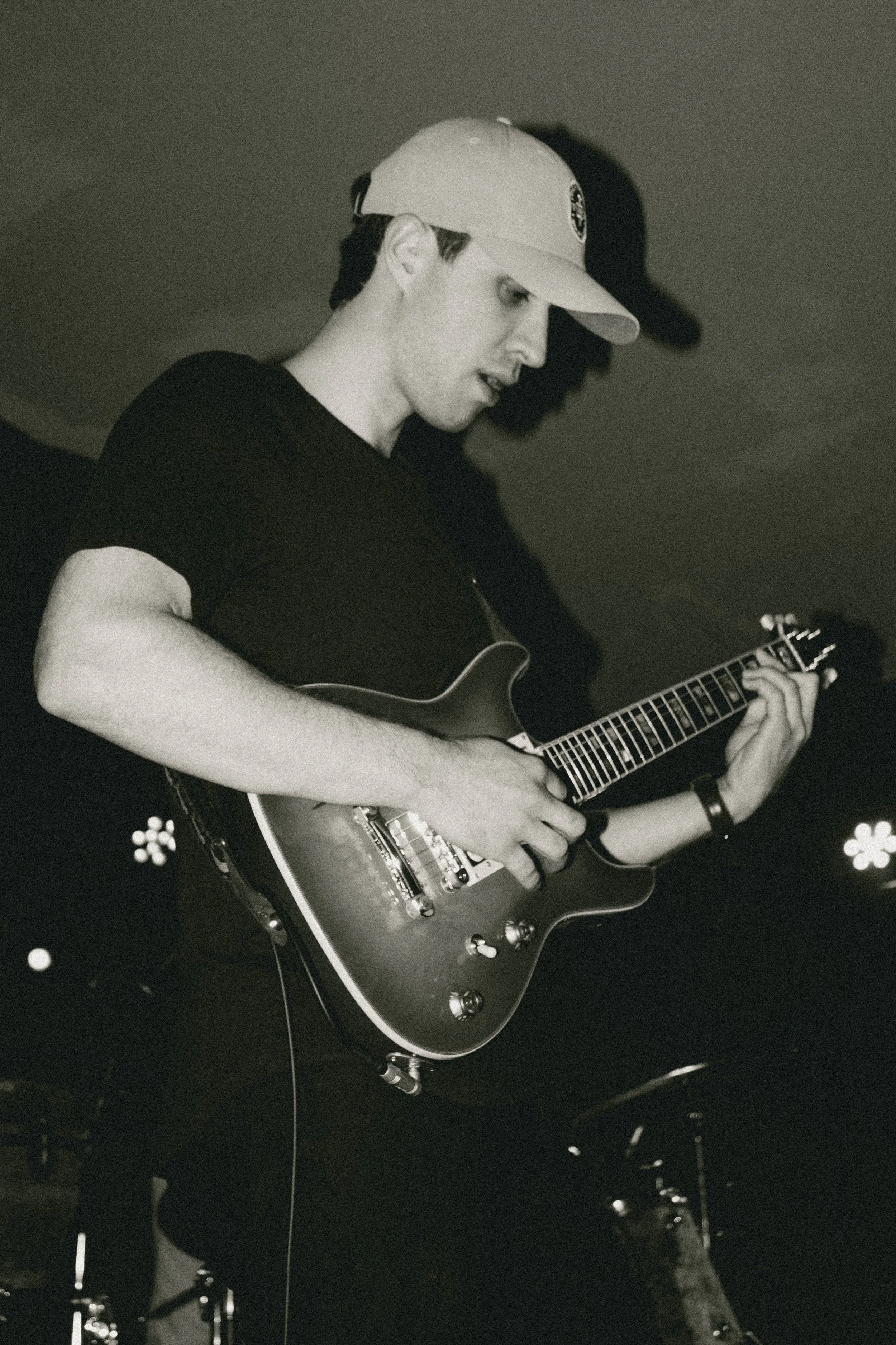 A young man playing an electric guitar while wearing a baseball cap, in a dimly lit setting.