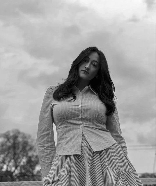 Black and white photo of a young woman standing outdoors with clouds in the sky and trees in the background. She has long dark hair, wearing a collared, striped button-up shirt and a striped skirt.
