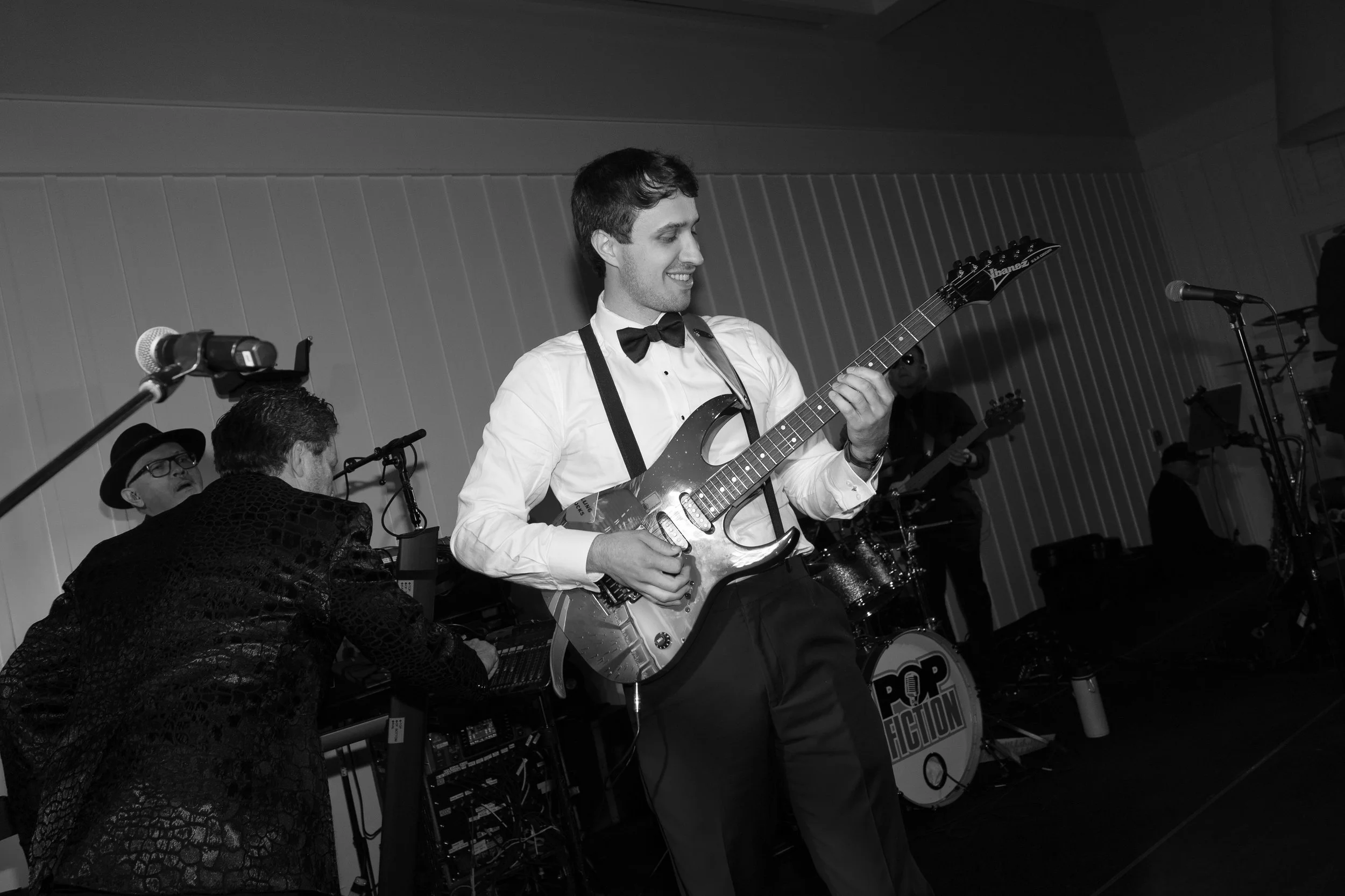 A young man in a white shirt and black bow tie playing an electric guitar on stage, smiling, with band members and musical equipment around him, at a live music event.