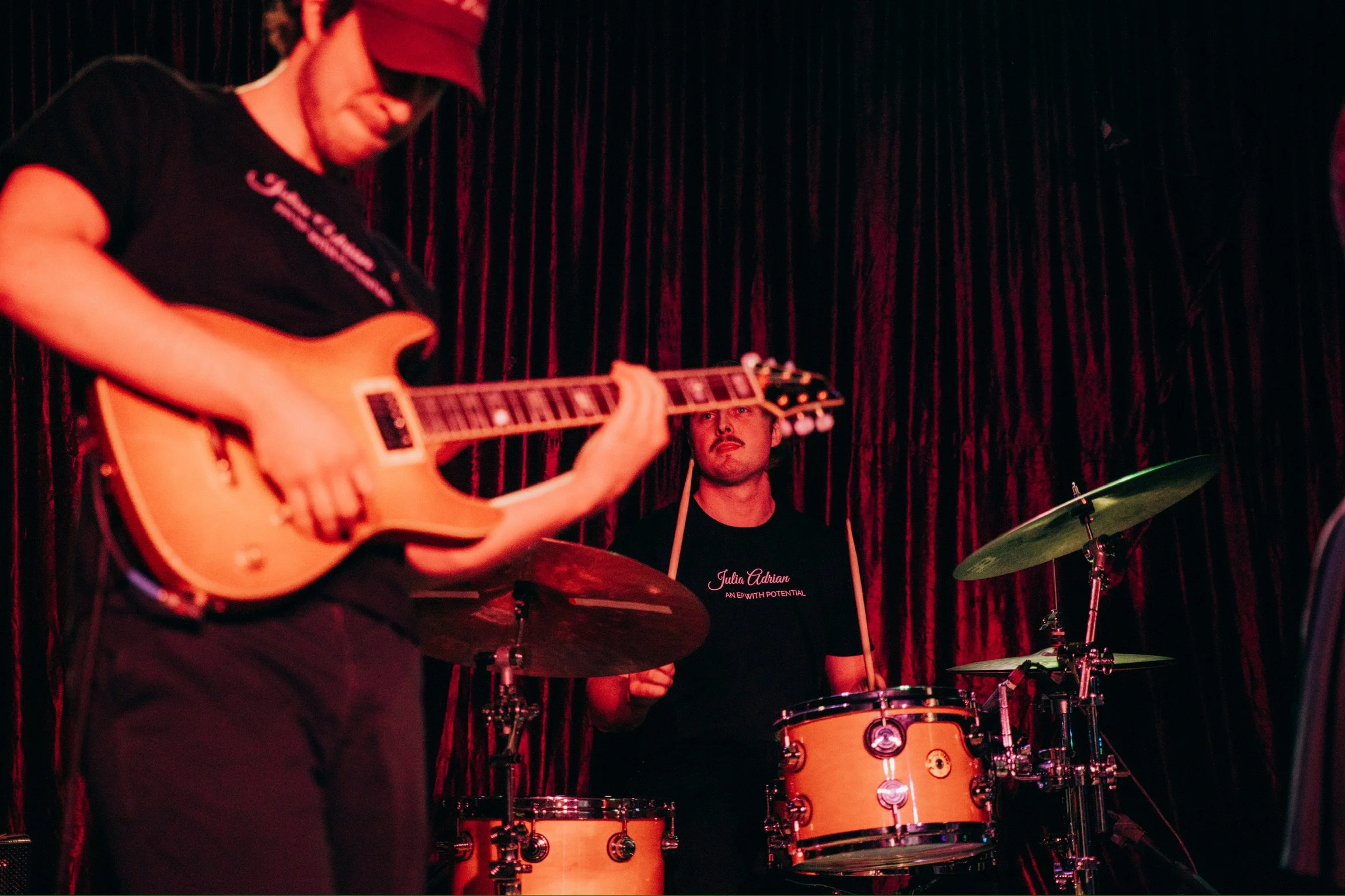 Musicians performing on stage with a guitarist in the foreground and a drummer in the background, set against a dark curtain with red lighting.