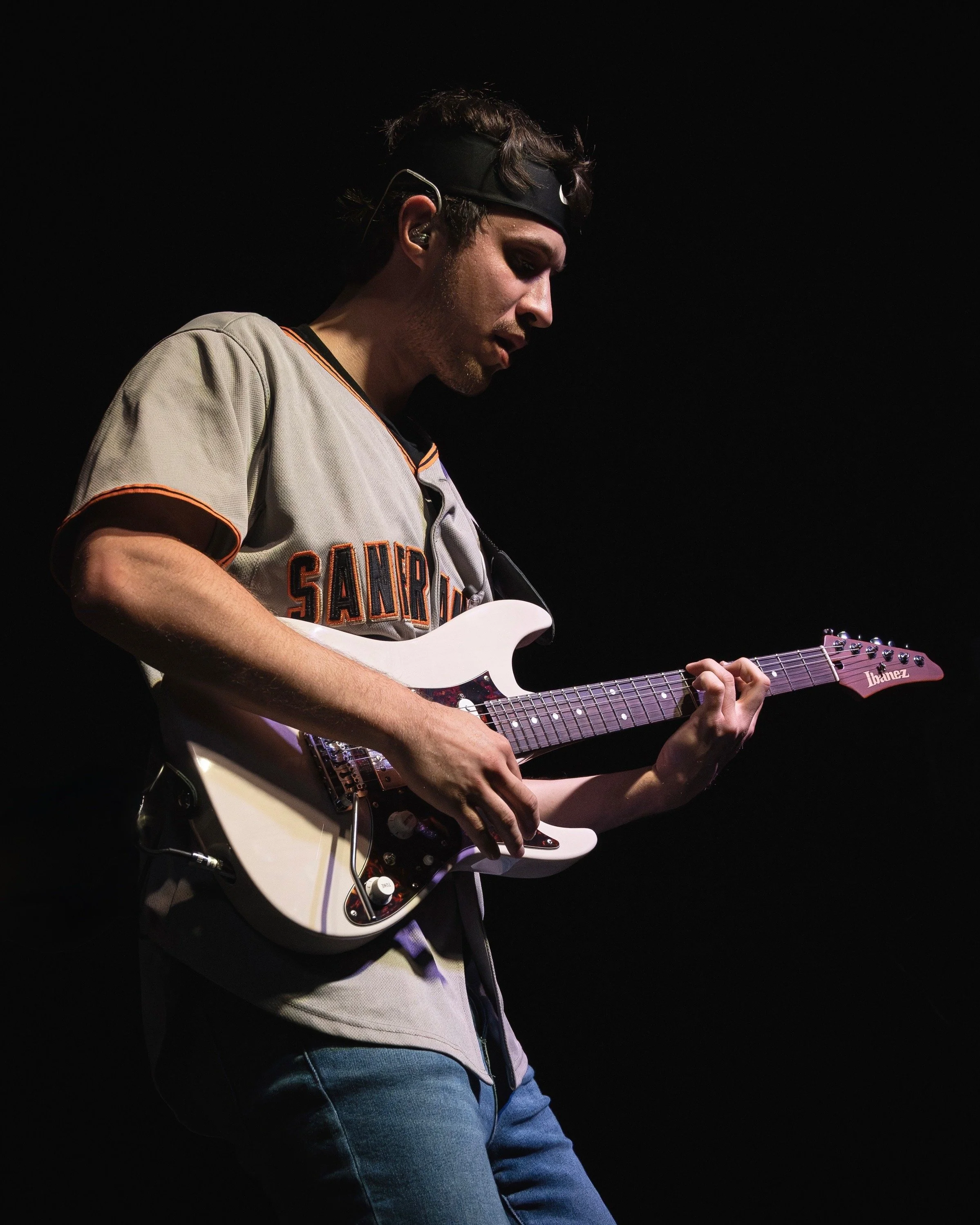 A musician playing an electric guitar on stage, wearing a San Francisco Giants baseball jersey and a black headband, with a dark background.