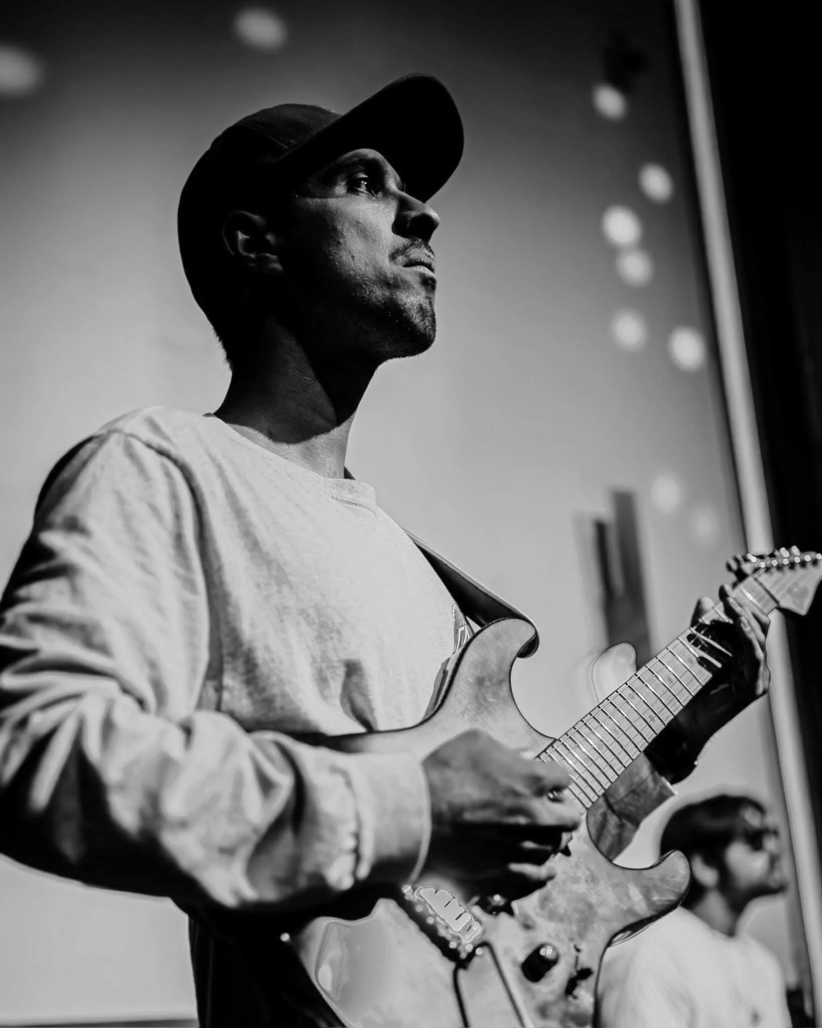 A black and white photo of a person wearing a cap and playing an electric guitar, with a blurred background and circular lights on the side.  Nashville Guitar Lessons Zoom Guitar Lessons Virtual Lessons In Person Guitar Lessons Session Work Audition