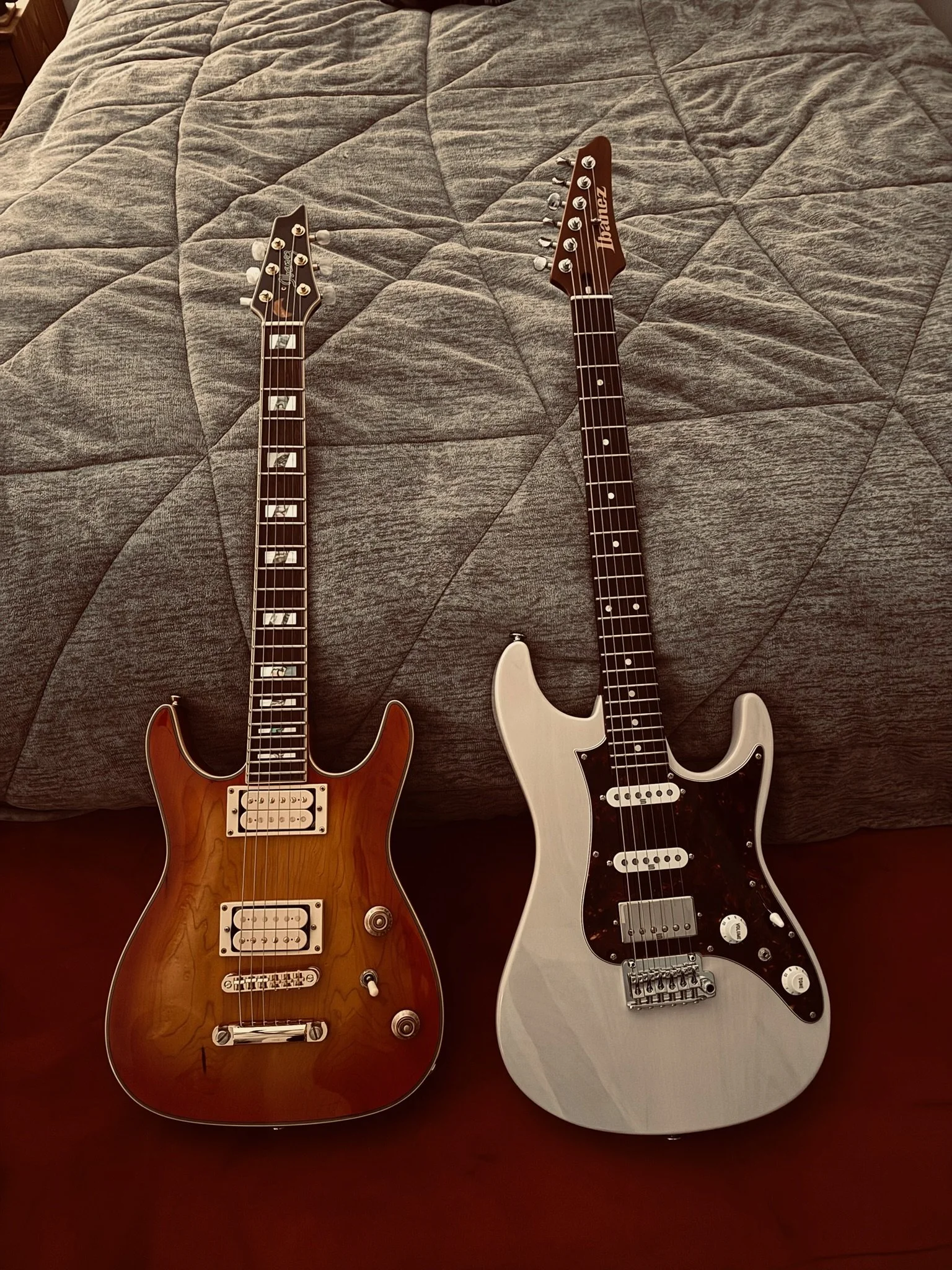 Two electric guitars lying on a bed with a gray quilted bedspread. One guitar has a natural wood finish, and the other is white.