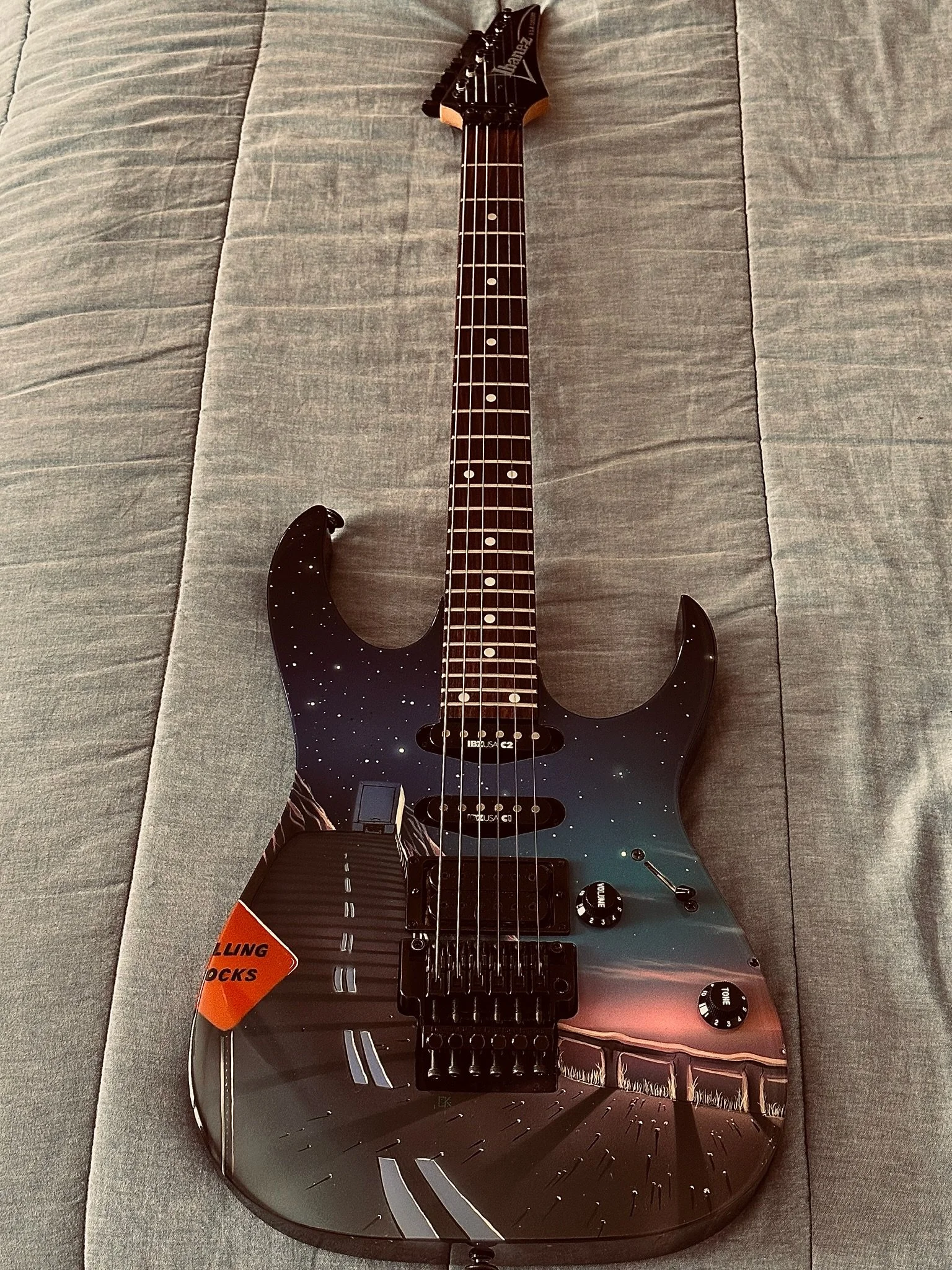 An electric guitar with a black finish and starry night design, featuring a sticker that says "HARD LANDING ROCKS" on the body, resting on a grey fabric surface.