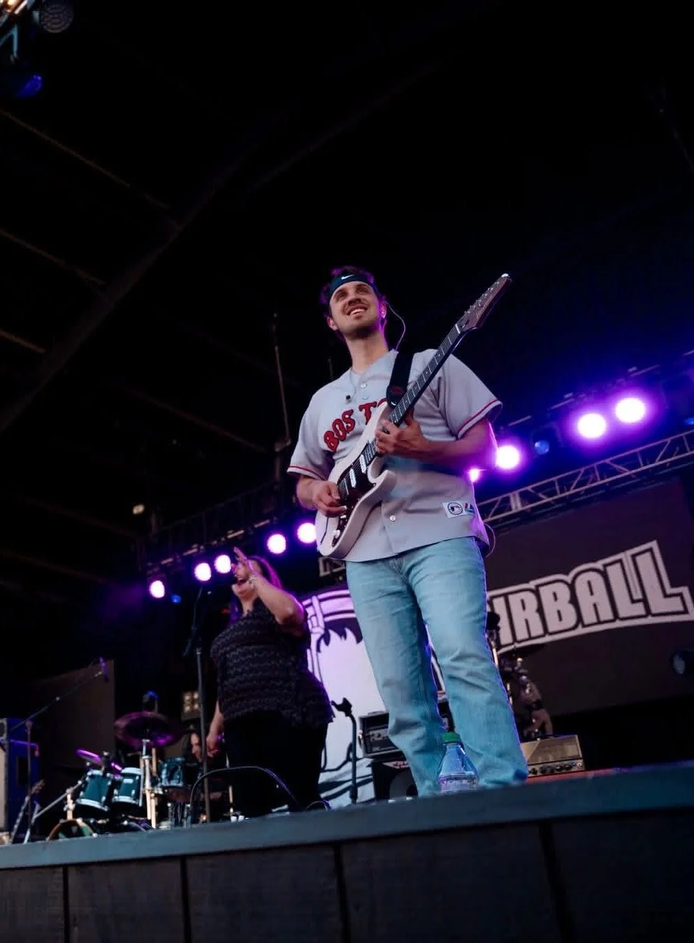 Musician playing guitar on stage with a woman singing into microphone behind him, stage lights in the background, and a banner reading 'FOOTBALL'.