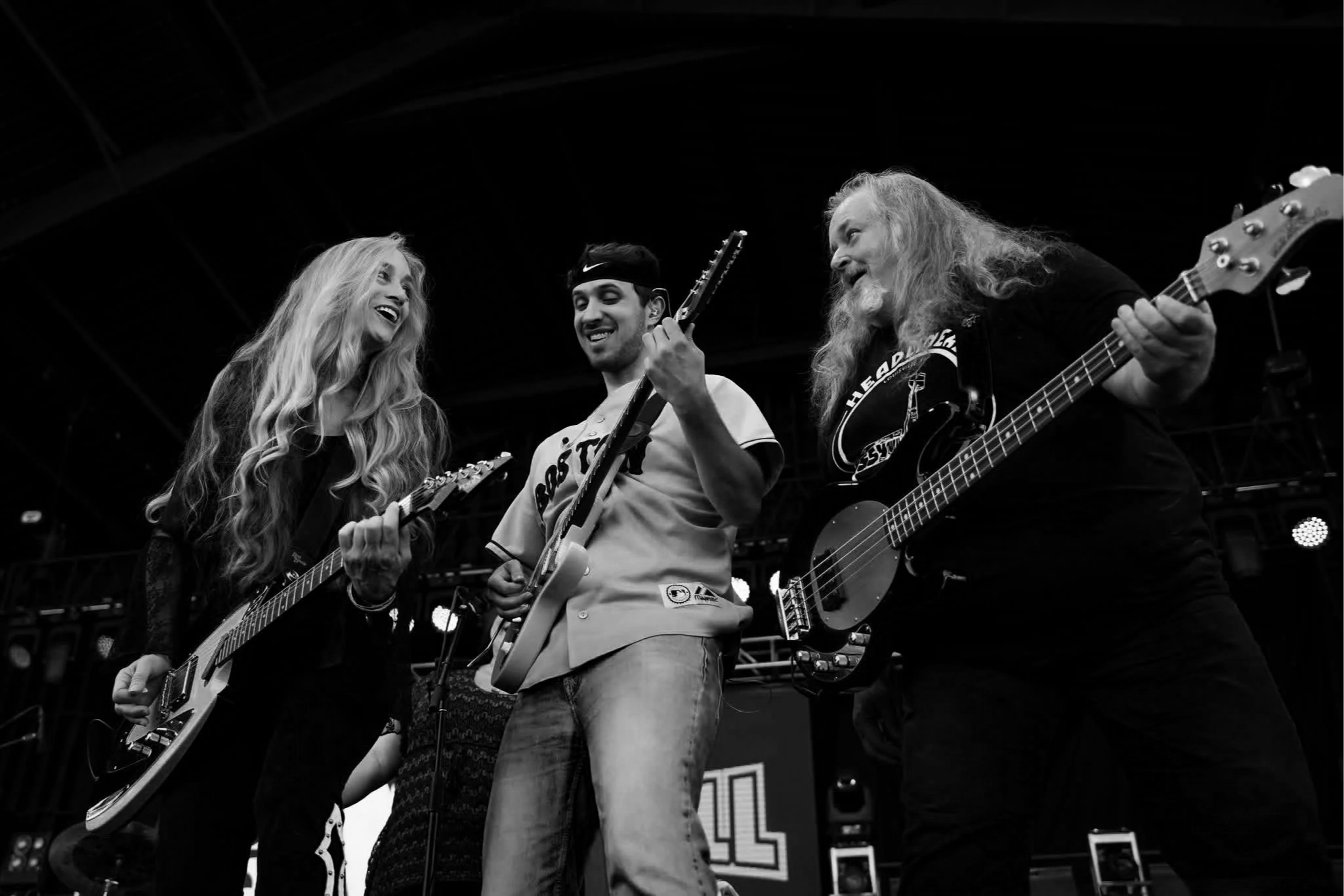 Three musicians performing on stage, two women and one man, playing guitars and singing, smiling and enjoying the moment, with a dark background and stage lighting.