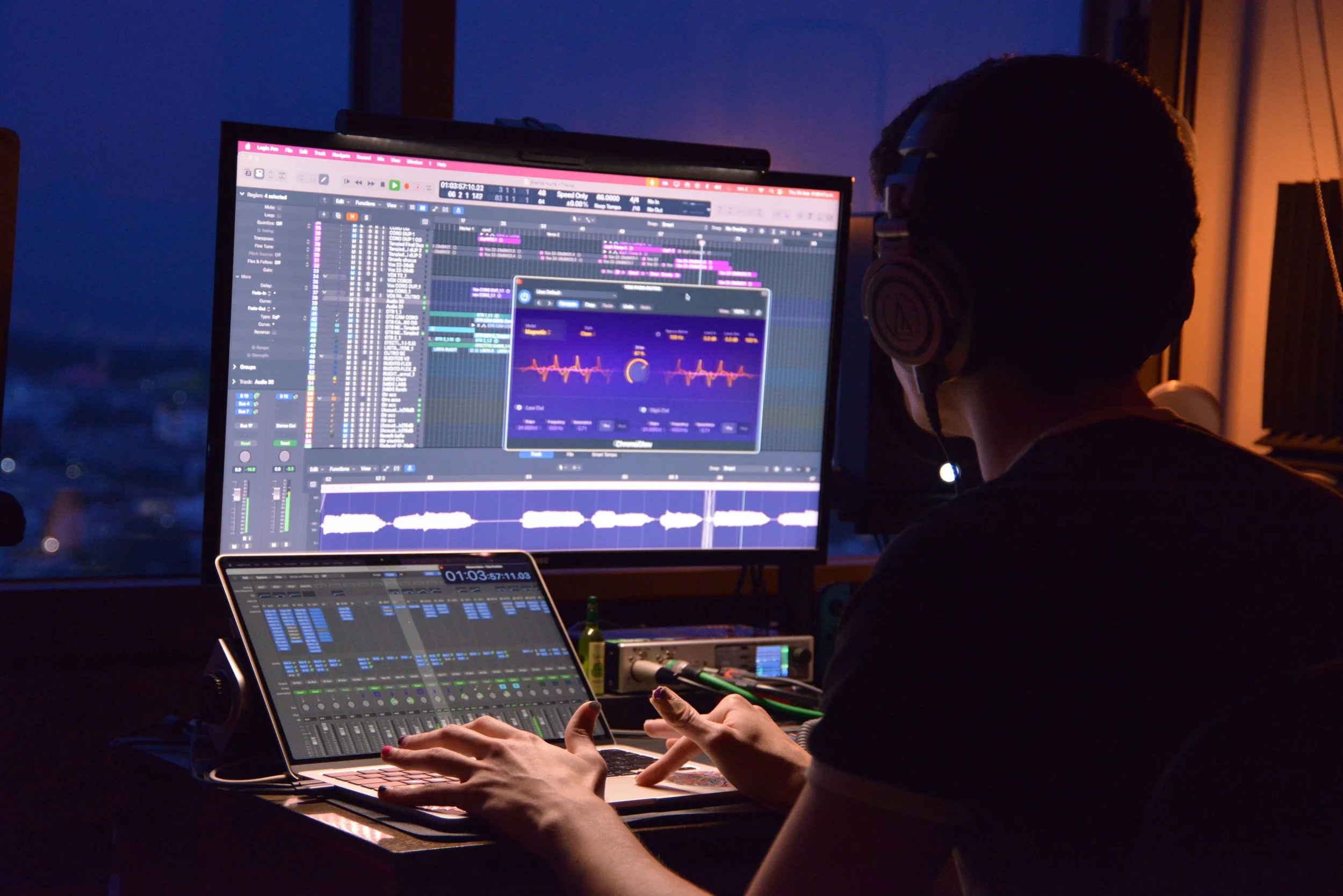 A person in headphones working on music production software on a computer, with a second screen and tablet displaying audio editing and waveform visualizations in a dimly lit room.
