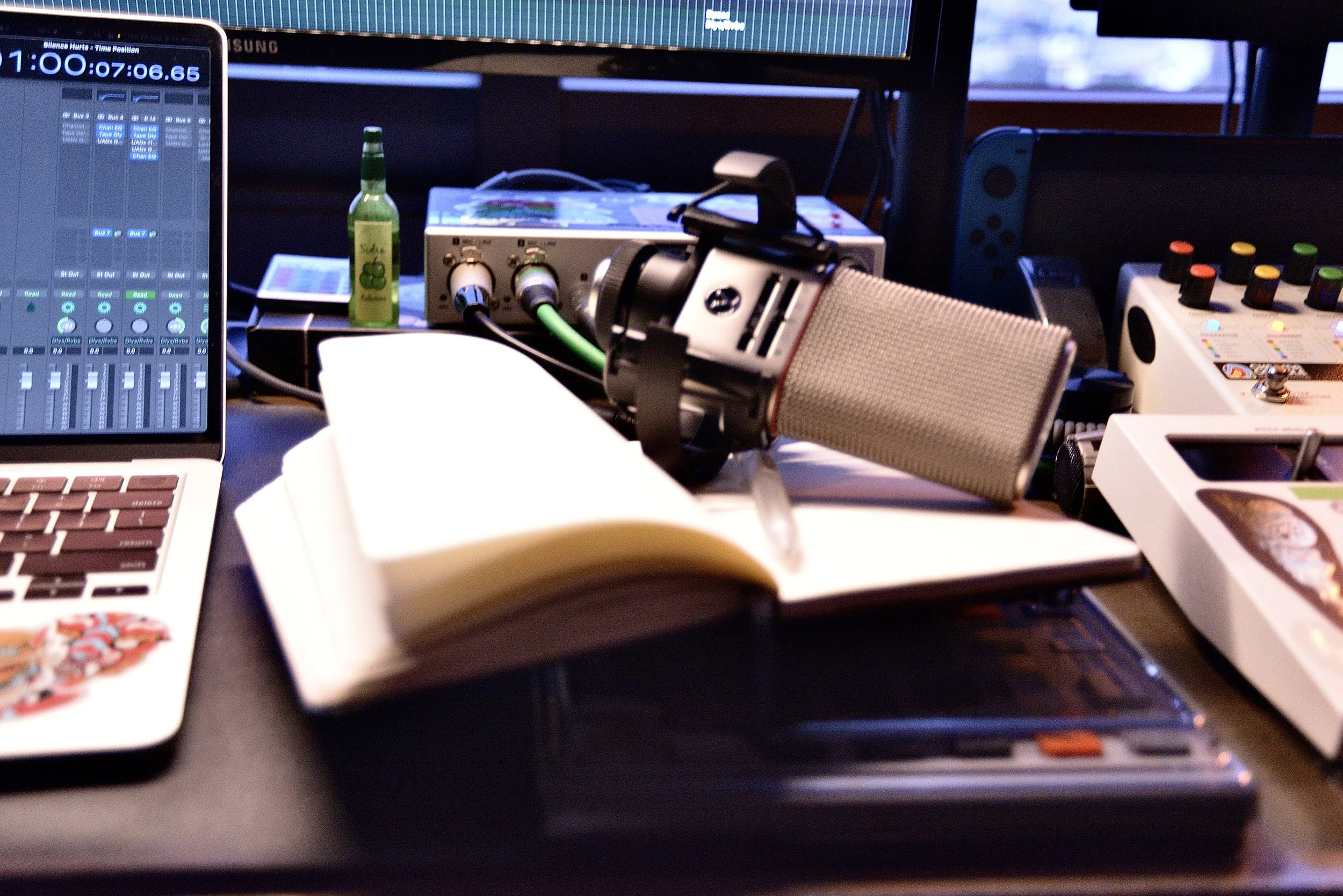 A cluttered workspace featuring a laptop with a digital audio work station, a large microphone, audio equipment and various papers and electronic devices.