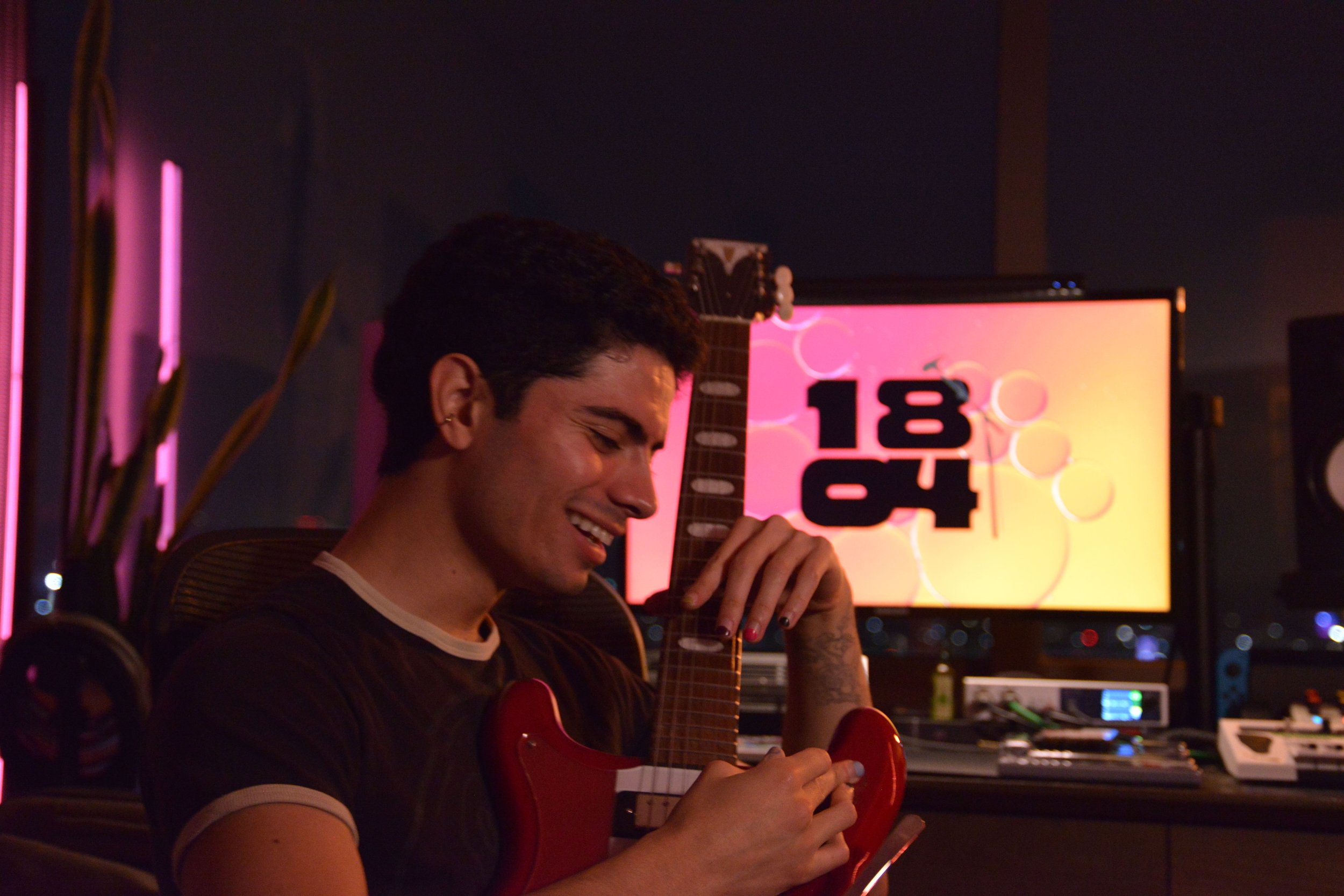 A young man with dark hair smiling and holding a red electric guitar in a dimly lit room with orange and pink neon lights and a digital display in the background reading 1804.