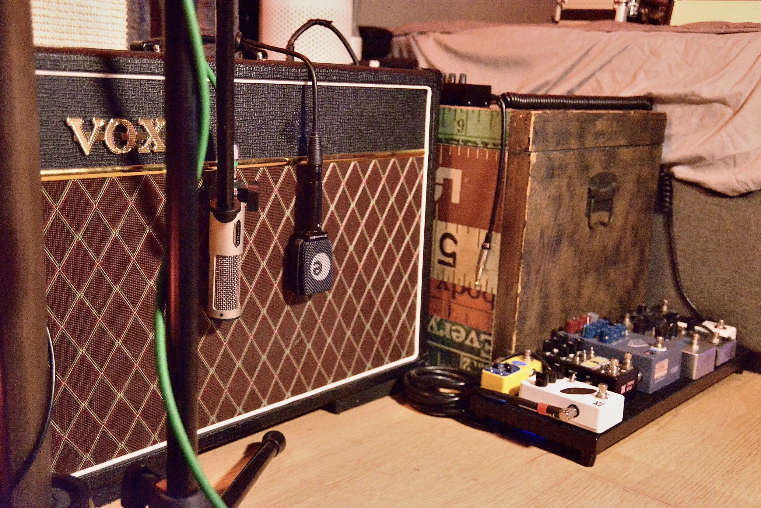 A collection of guitar amps, multiple guitar pedal effects, and various cables on a wooden floor in a music or rehearsal room.