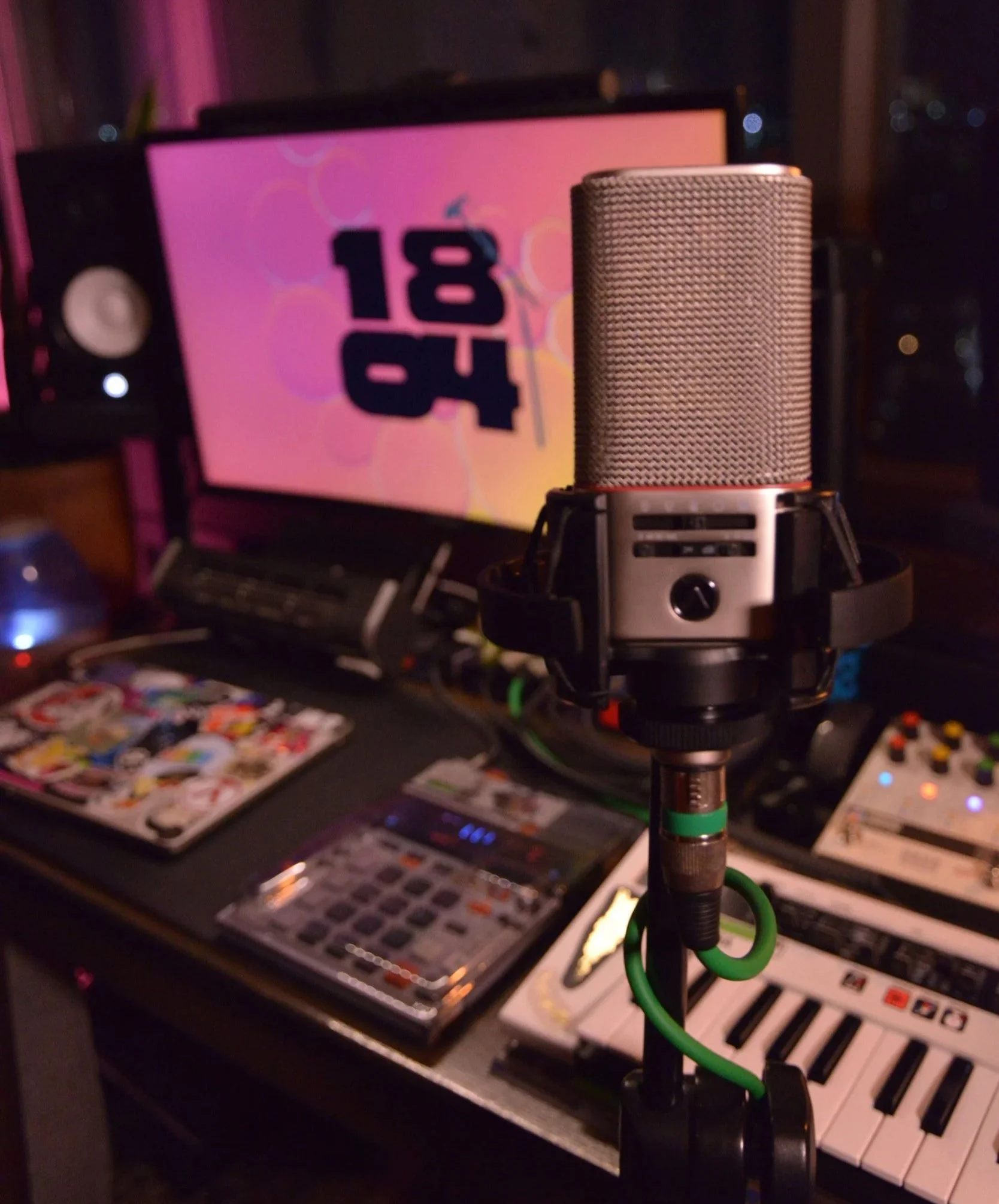 Studio microphone on a stand in front of a keyboard, with a music production setup including a computer monitor displaying '18' and '04' in the background.